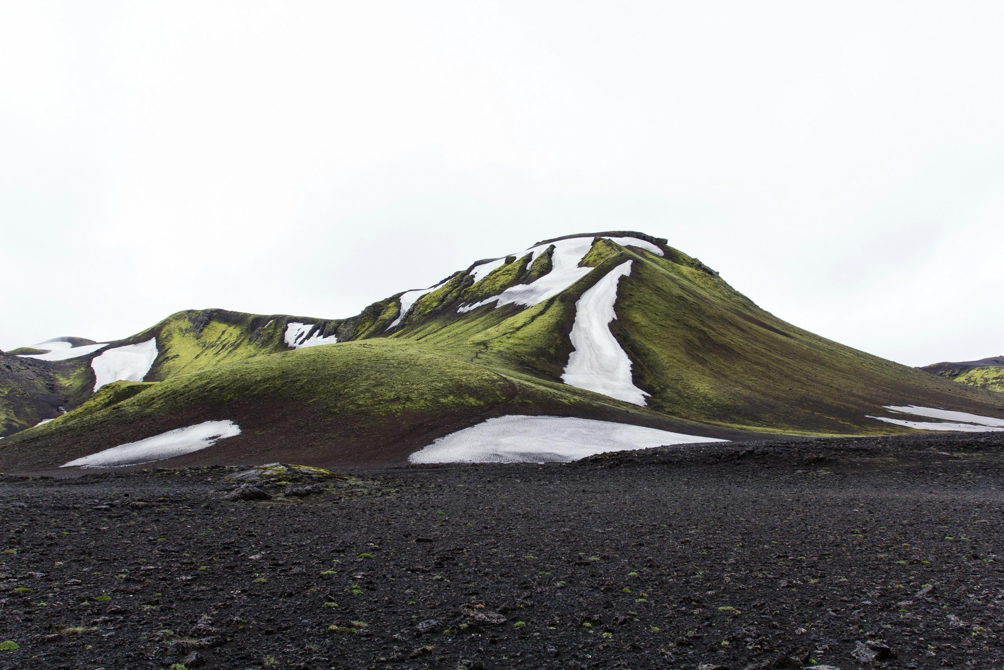 Green moss-covered hill with patches of snow set against a stark black volcanic landscape in the Icelandic Highlands.