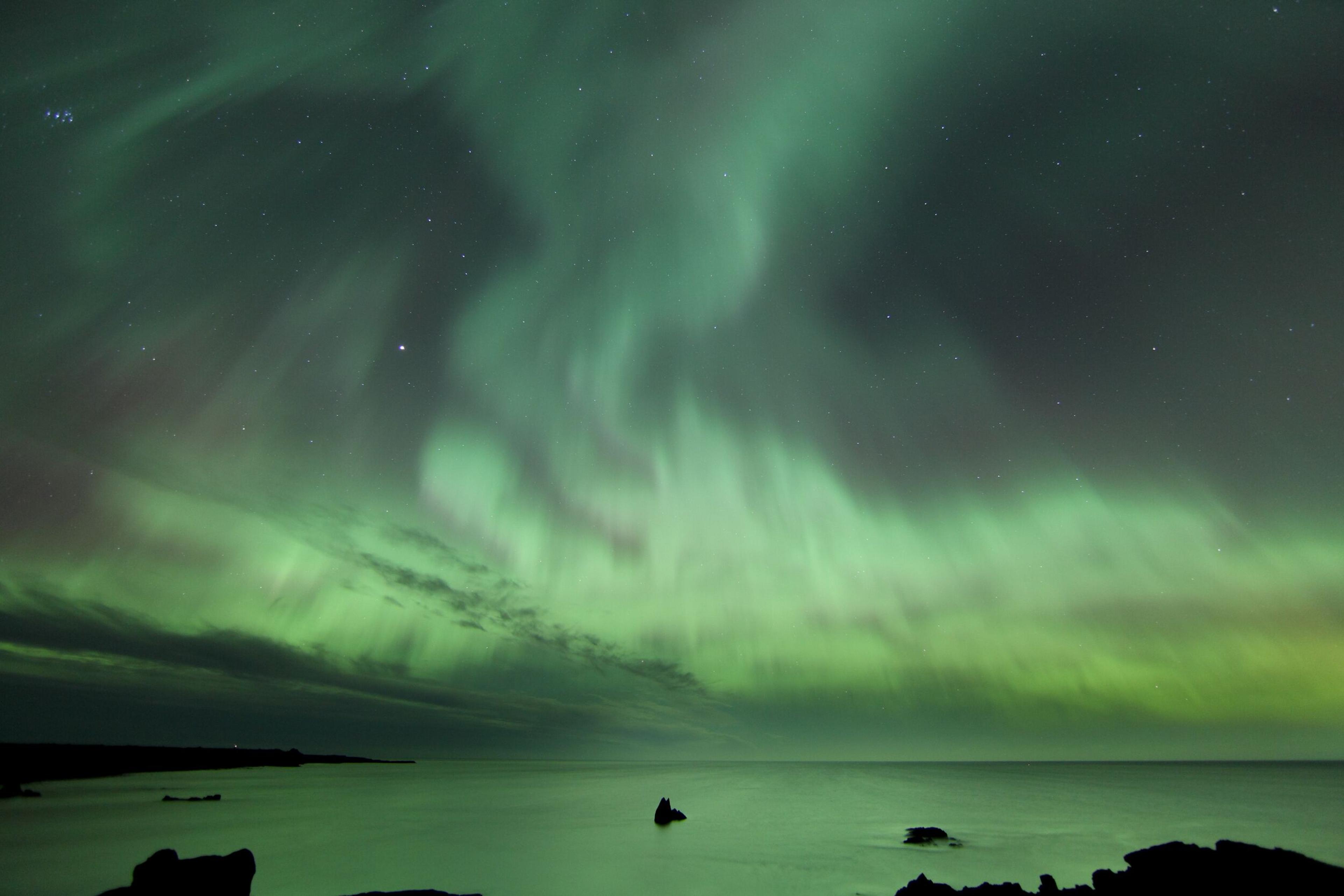 Vibrant green Northern Lights dance across the night sky, reflecting on the calm surface of the ocean.