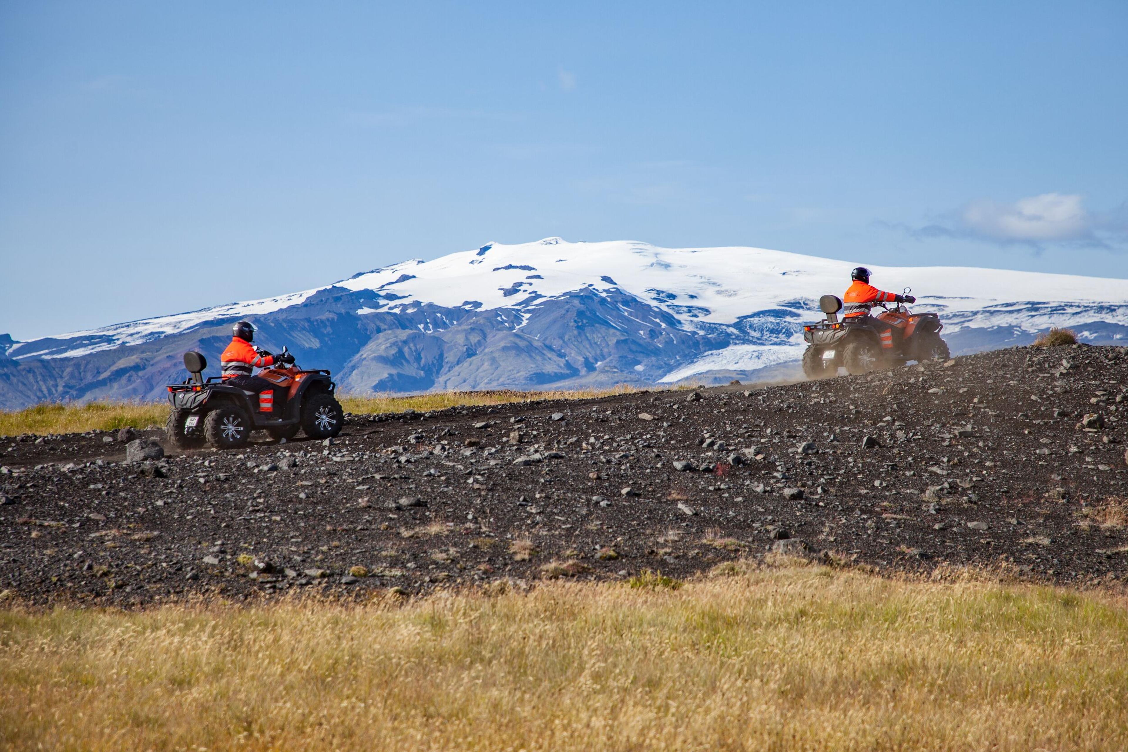 A group of ATV riders wearing bright orange jackets navigate a winding dirt trail through black sand dunes along the Icelandic coast, with Myrdalsjökul glacier in the background.