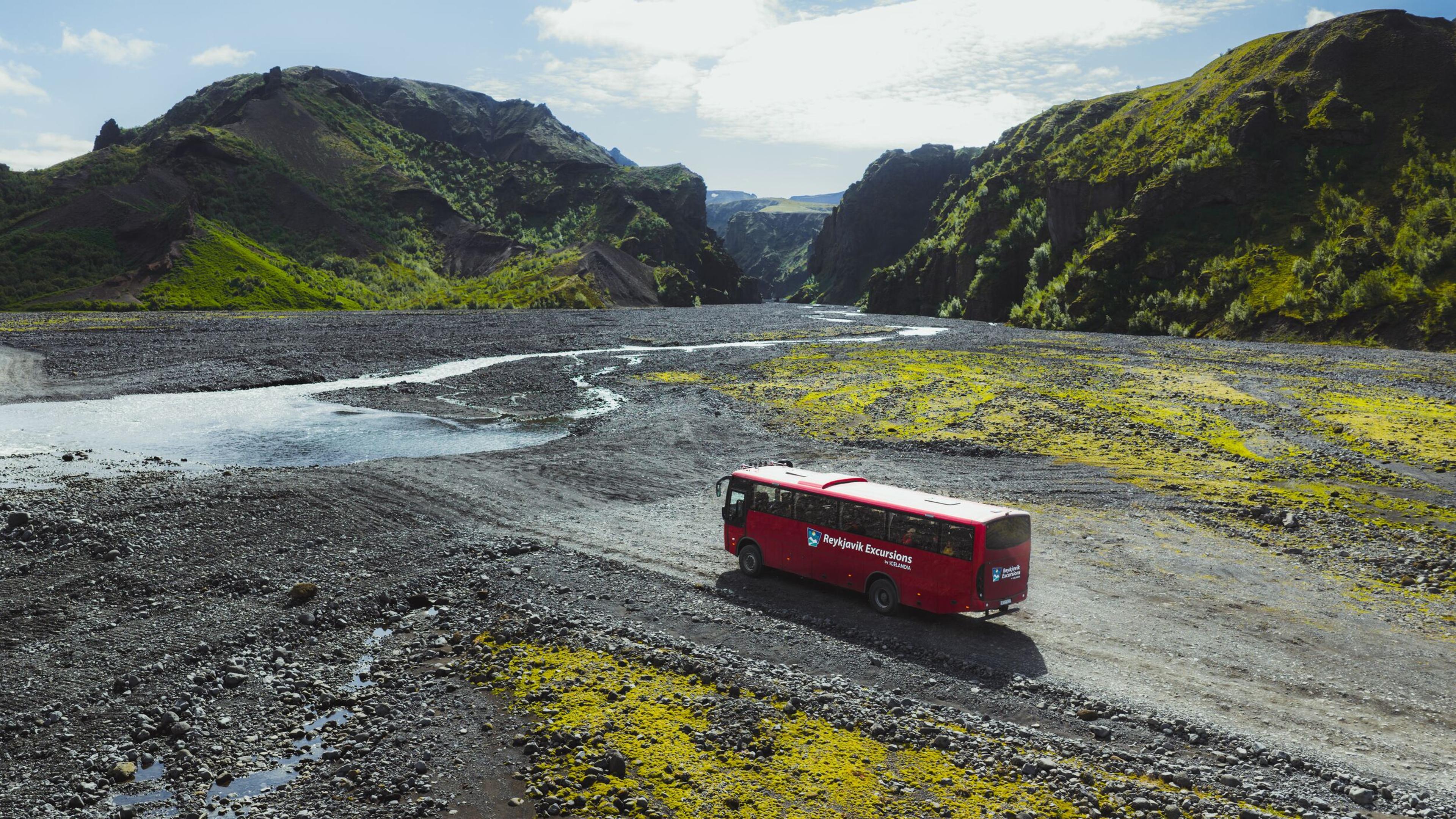 A red tour bus drives on a rocky riverbed through a scenic valley surrounded by green mountains.