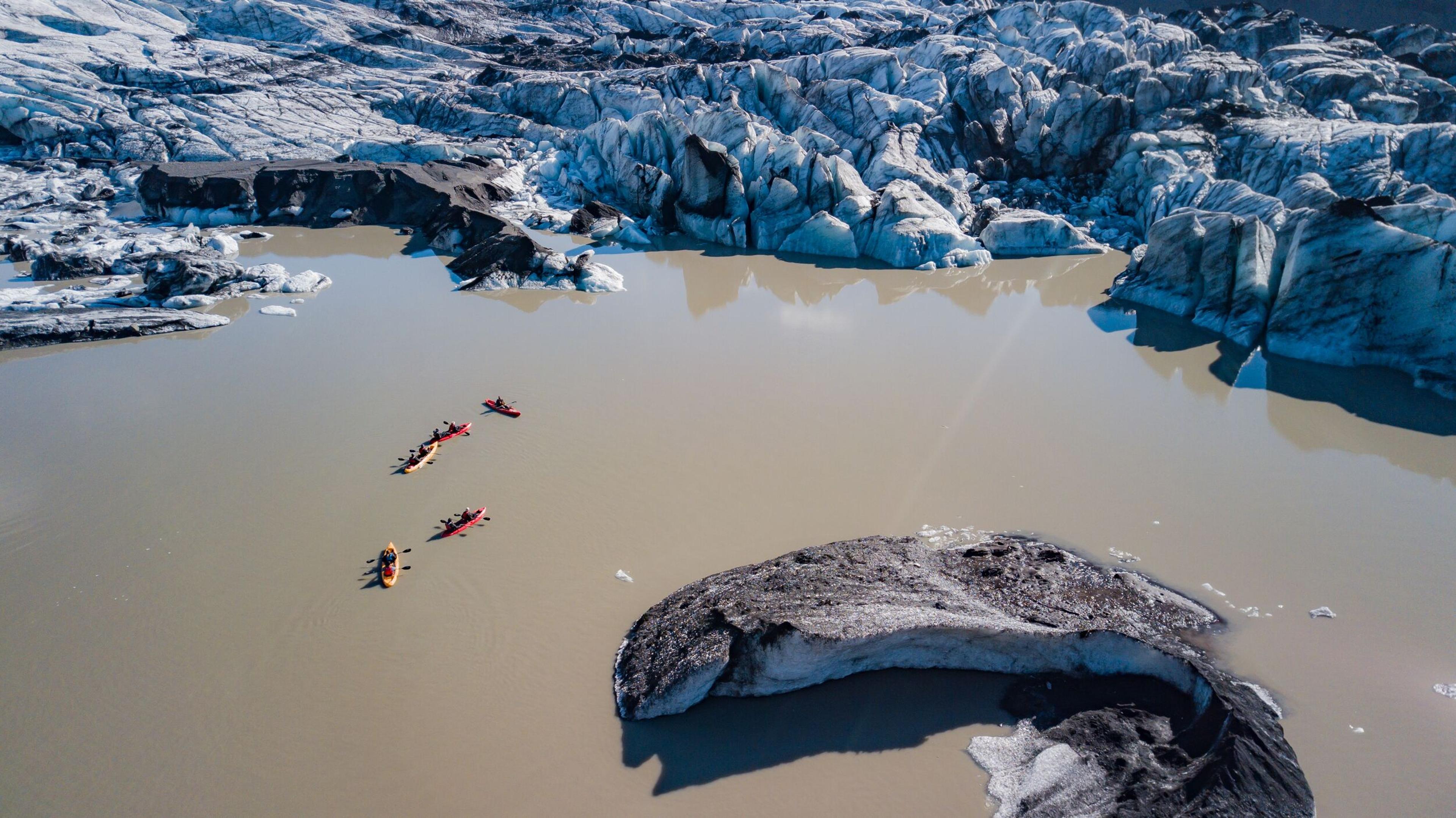 A group of kayakers in red and yellow kayaks navigating a glacial lagoon surrounded by rugged, ice-covered terrain, perfect for kayaking in Iceland.