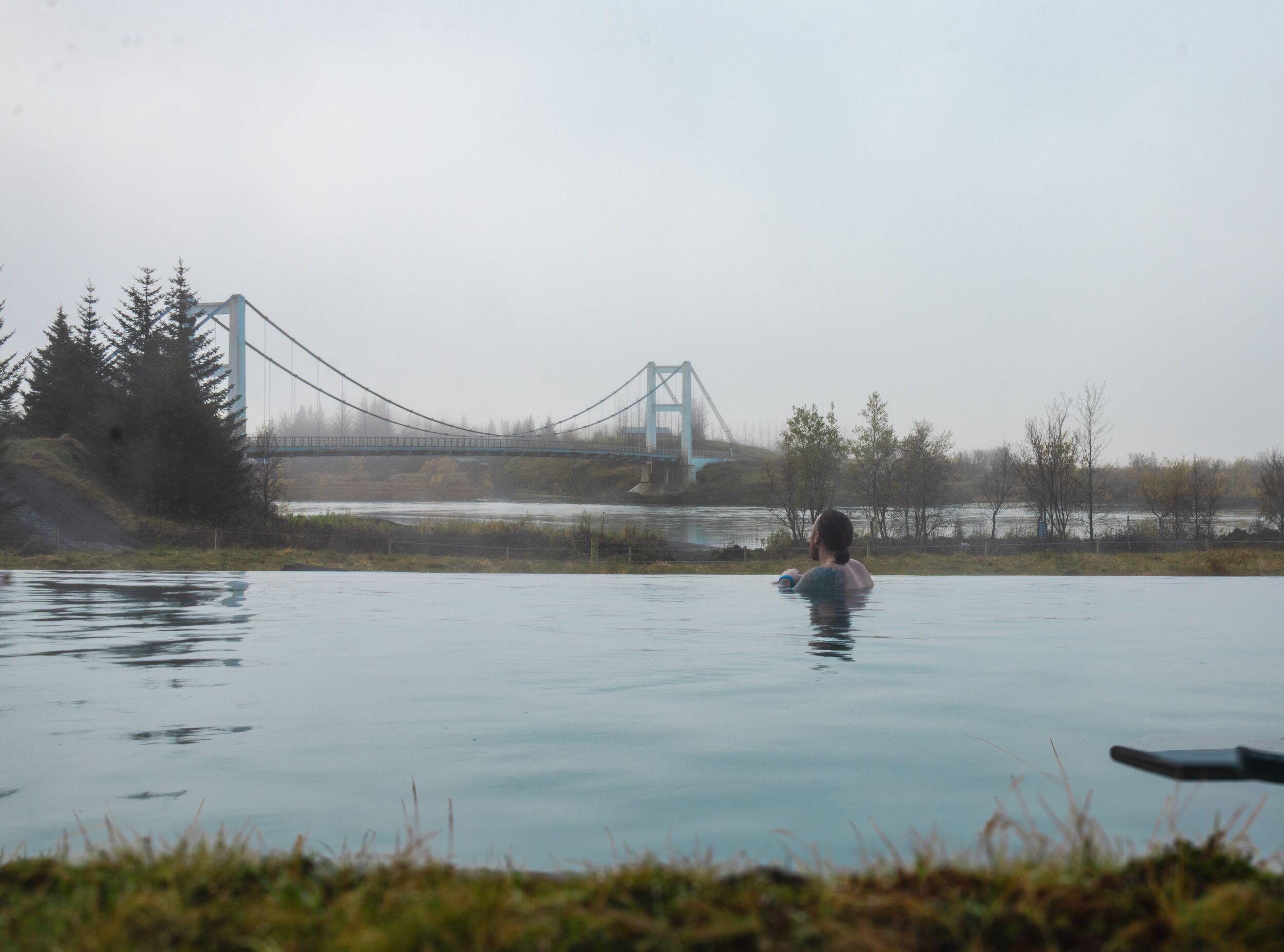 A person in an outdoor pool looks towards a suspension bridge on a misty day.