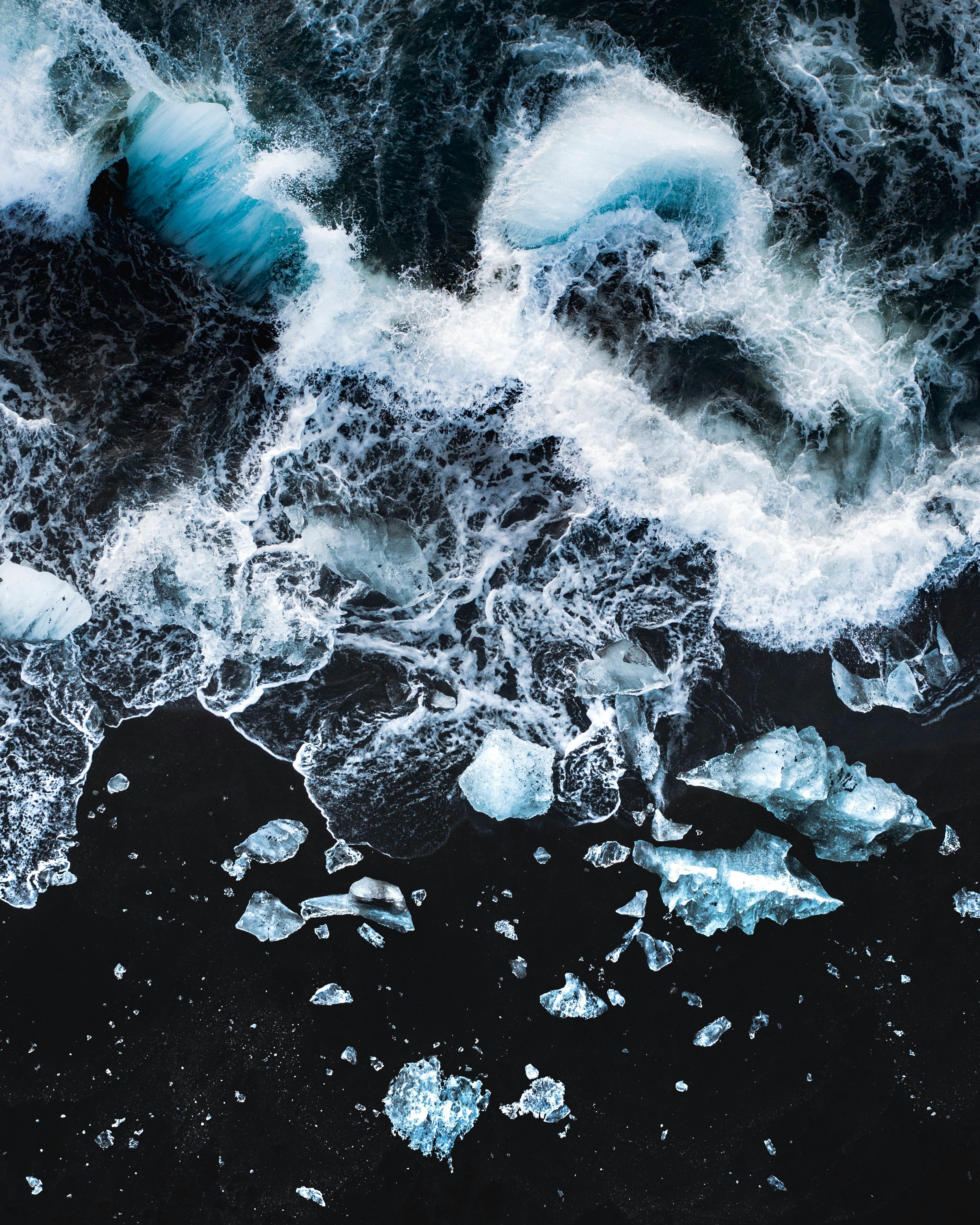 Aerial view of a dark shoreline with scattered pieces of blue-tinged ice, contrasting against the white foam of the breaking waves.
