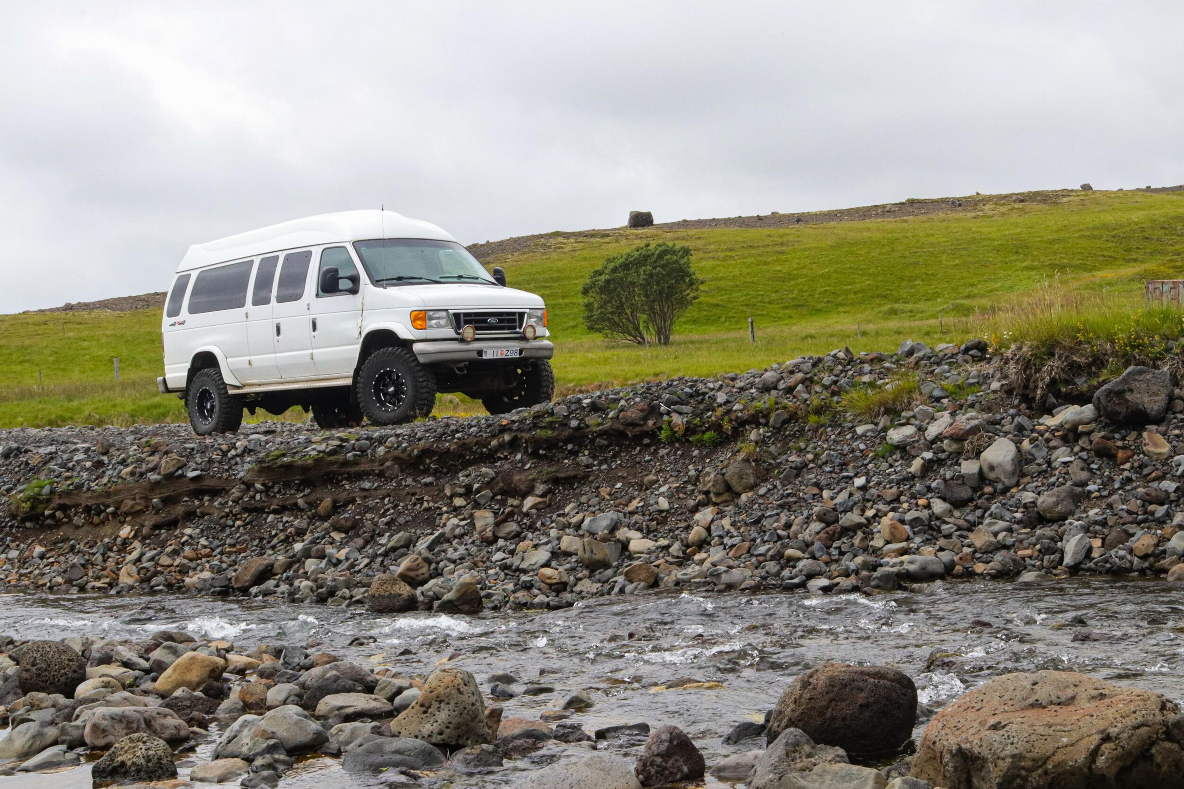 White off-road van driving on rocky terrain near a stream, with green hills in the background.