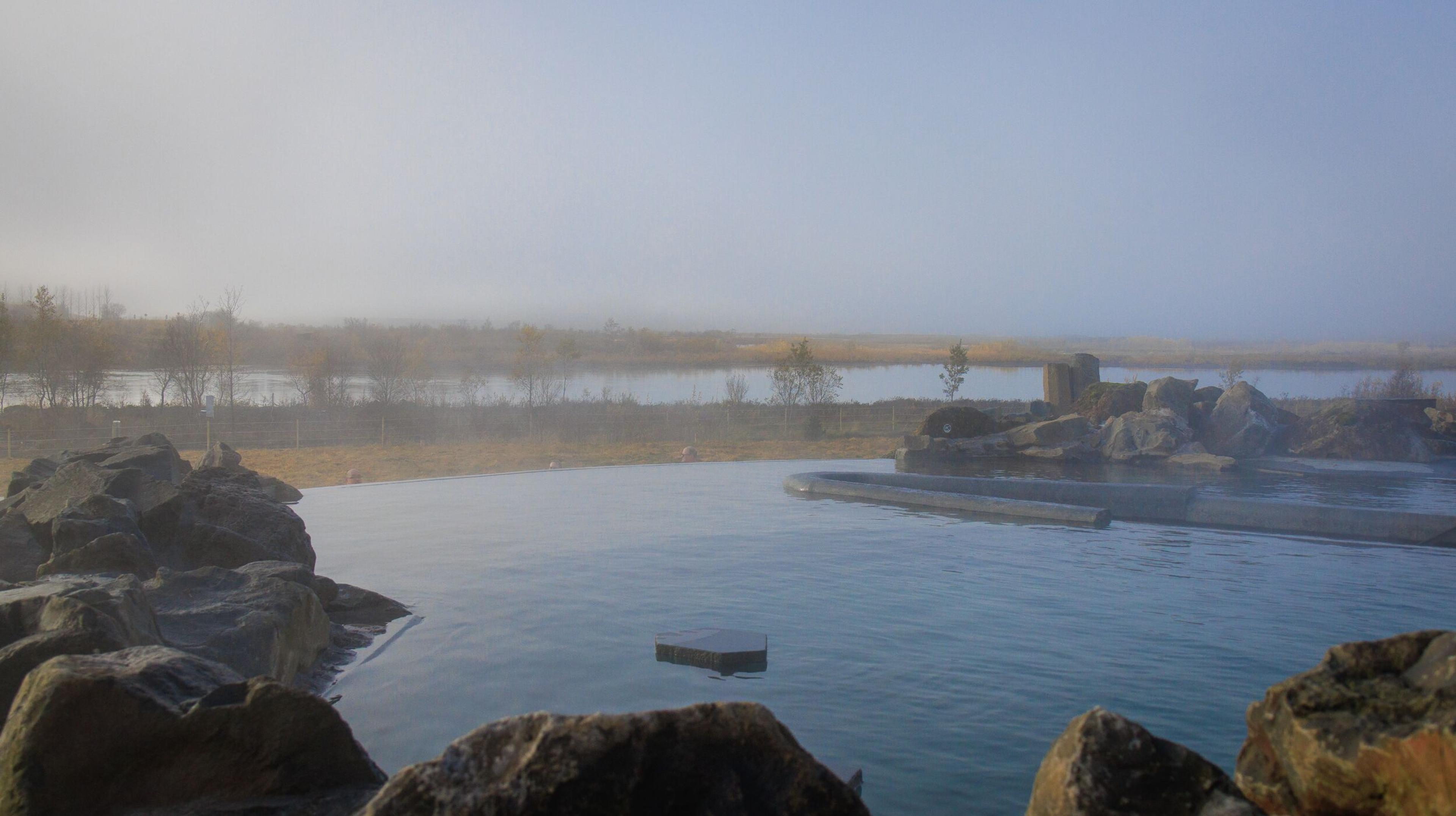 A steaming geothermal pool surrounded by rocks, with a misty lake and hazy sky in the background.