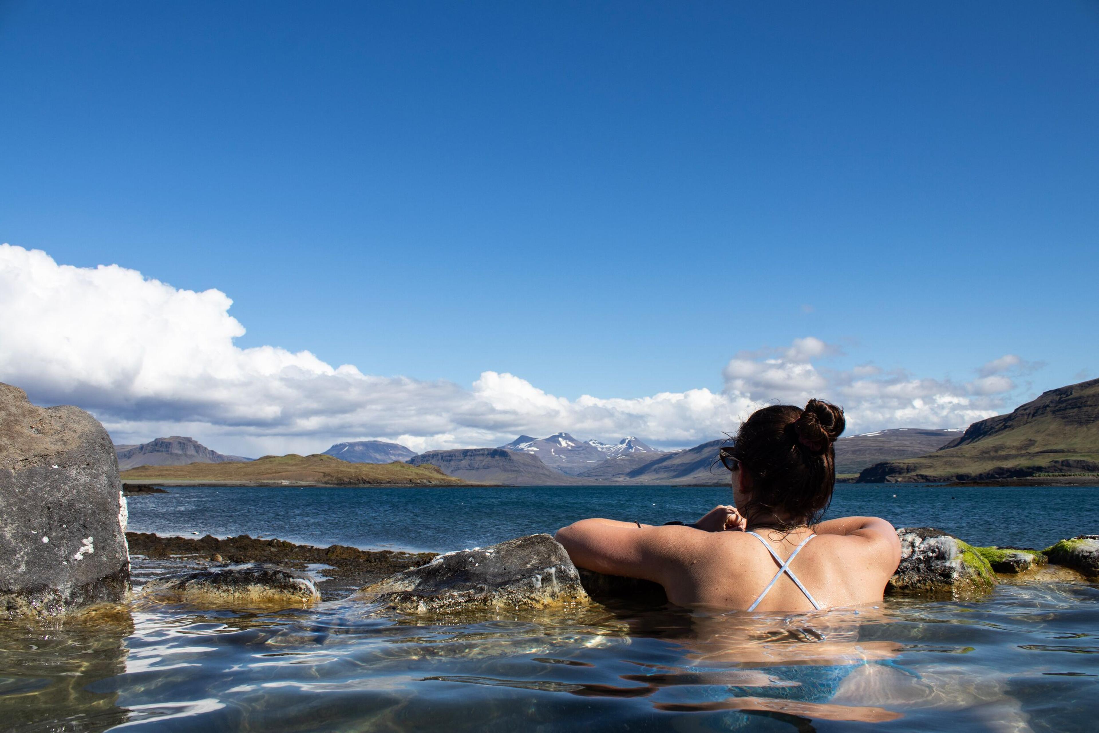 A person relaxes in a natural hot spring, looking out at a scenic bay with mountains under a blue sky.