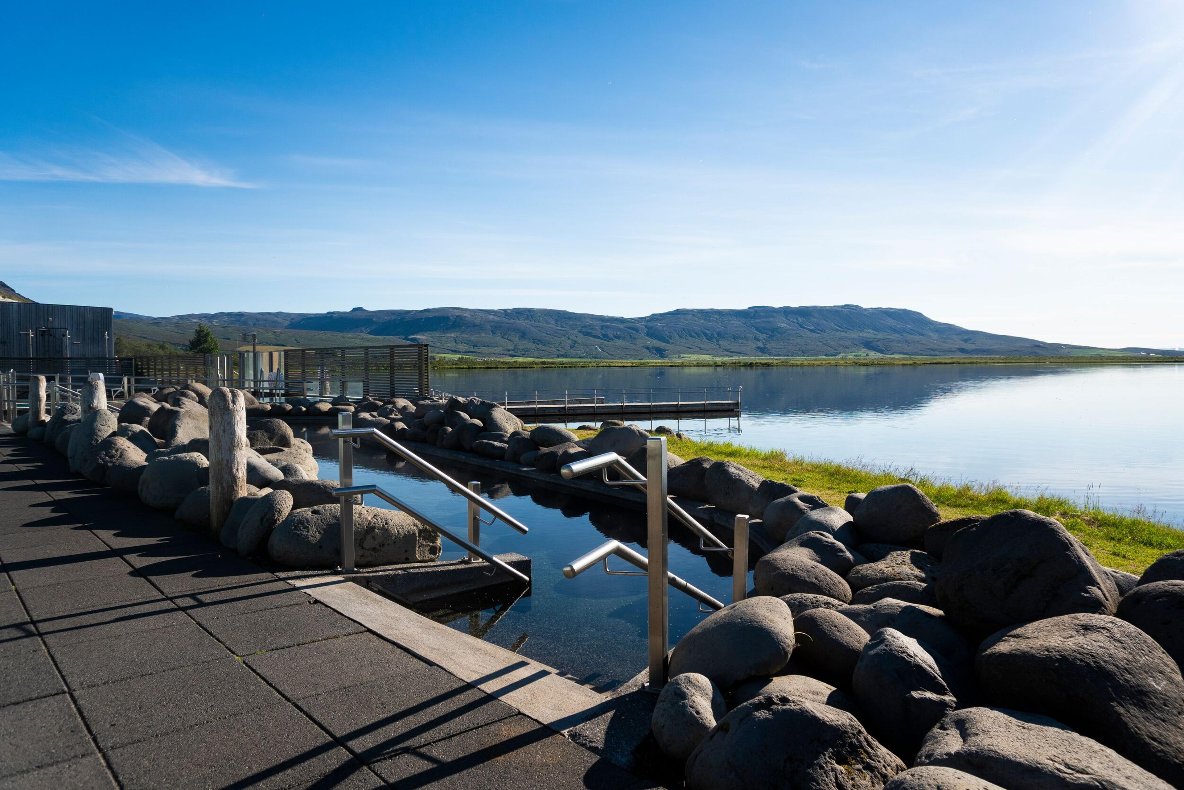An outdoor pool with metal handrails and dark rocks, beside a lake backed by mountains under a blue sky.