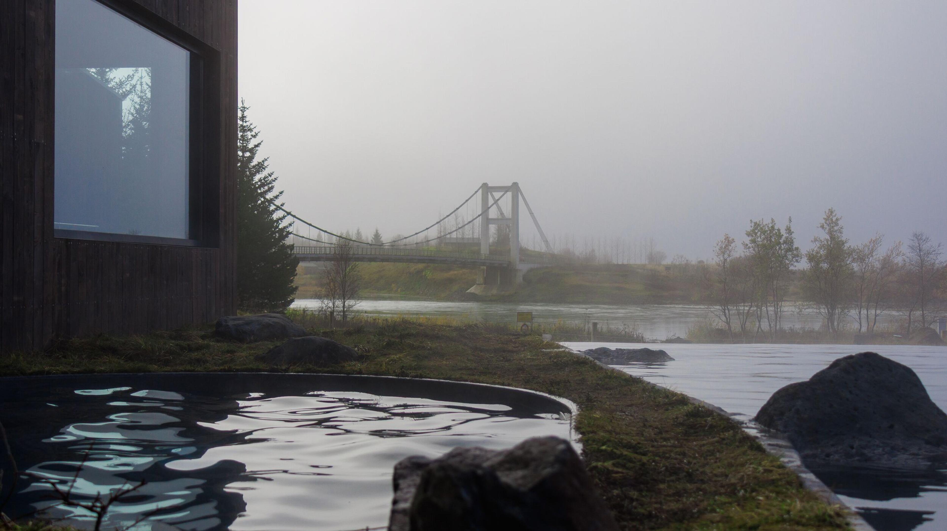 A hot tub sits outside a modern building, overlooking a misty river with a suspension bridge in the distance.