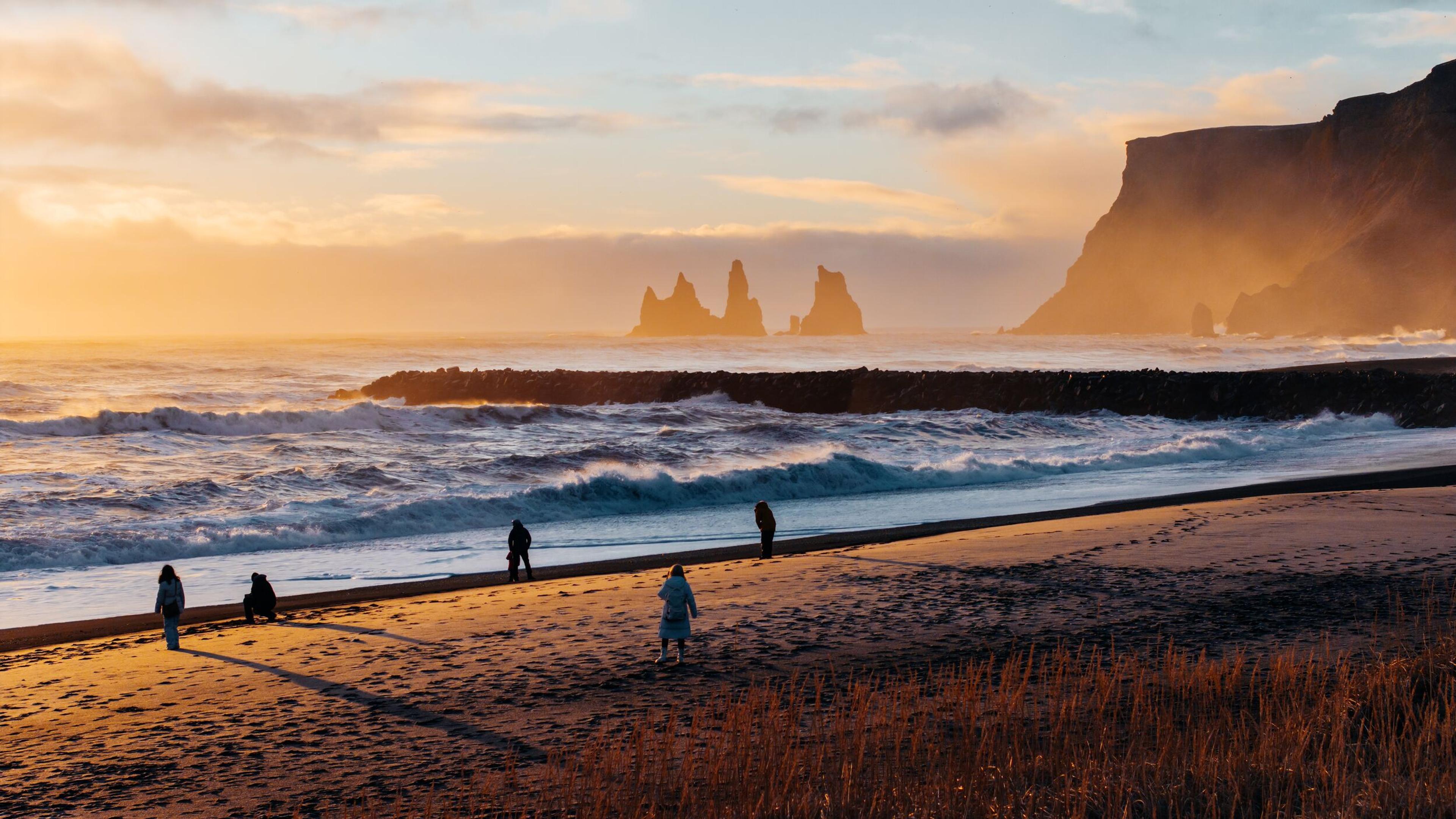 People on a black sand beach with crashing waves, sea stacks, and cliffs under a golden sky.