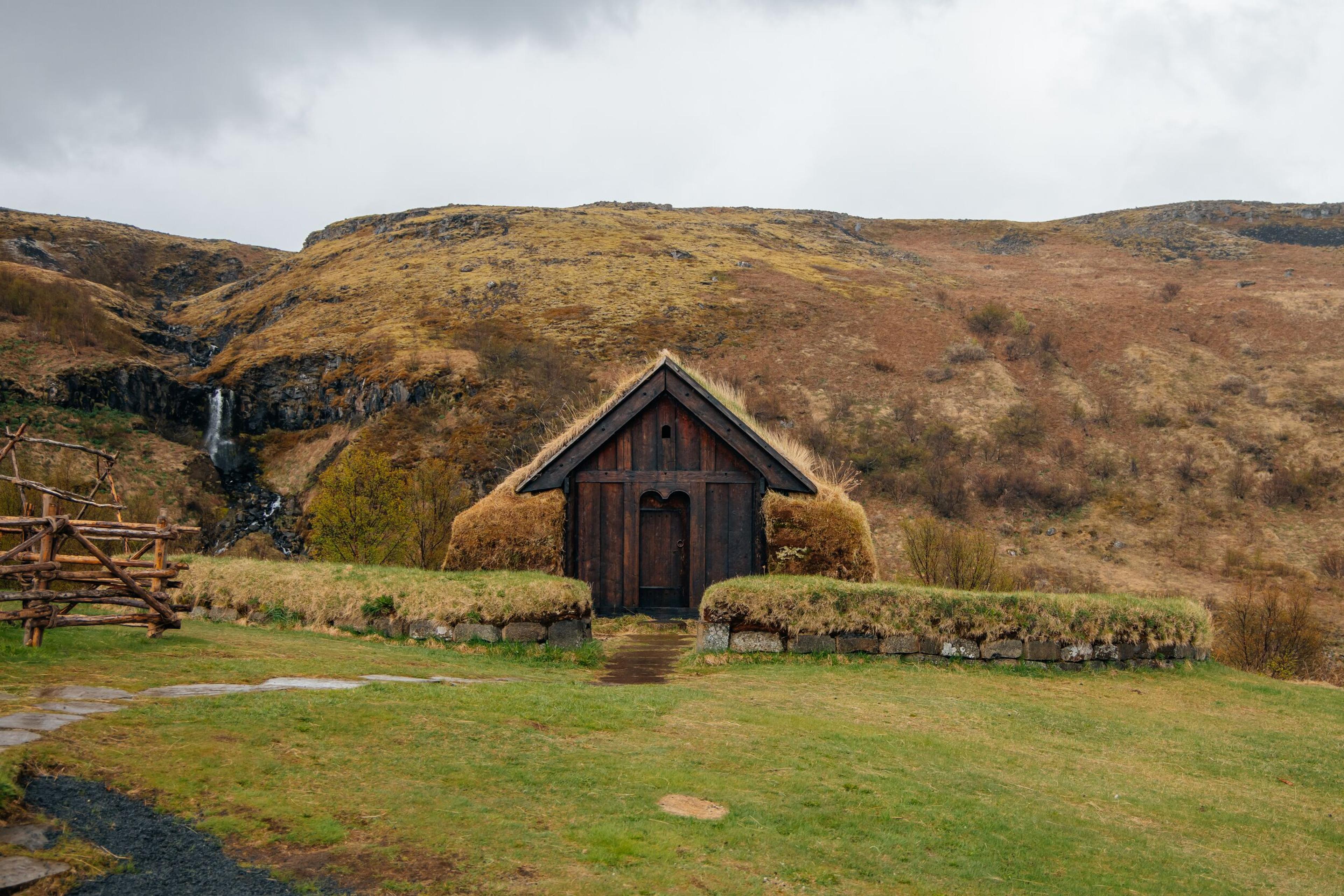 A traditional dark wooden turf house with a grass roof stands in a green field, backed by a brown mountain with a waterfall under an overcast sky.