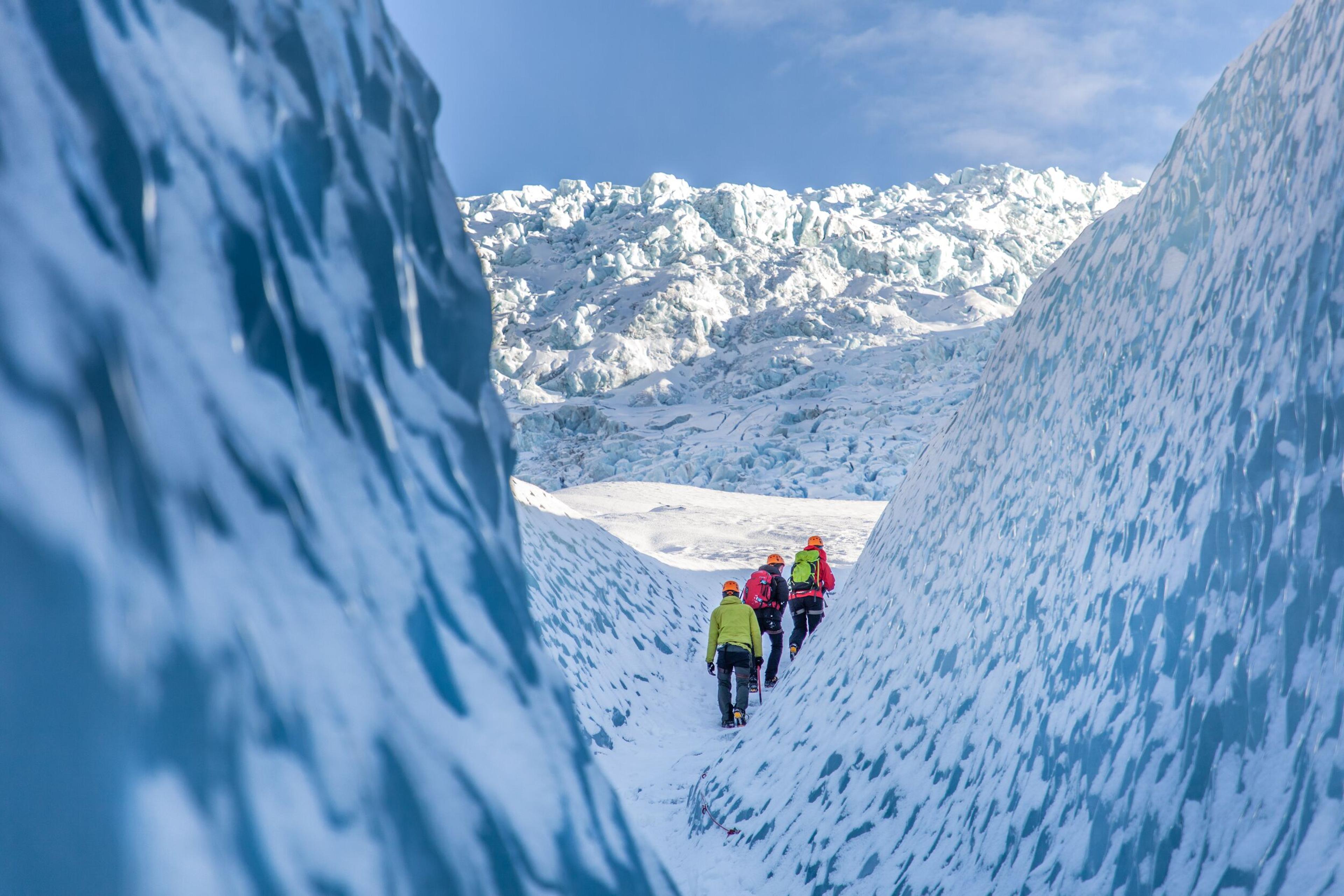 Three people hiking on a glacier, photographed from between two blue ice walls