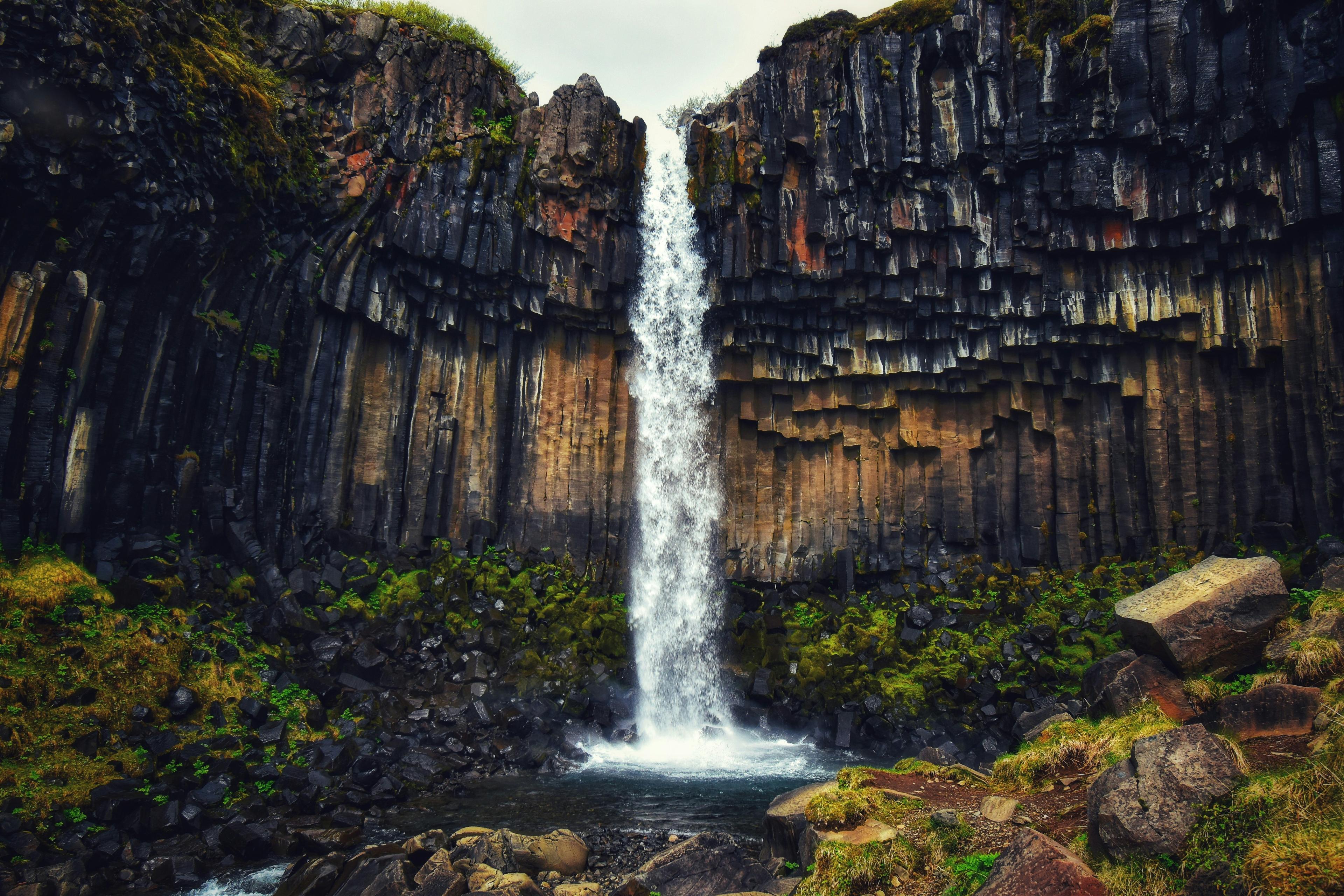 A majestic waterfall descending from a height, framed by striking columnar basalt formations, with lush greenery and rocks at its base.