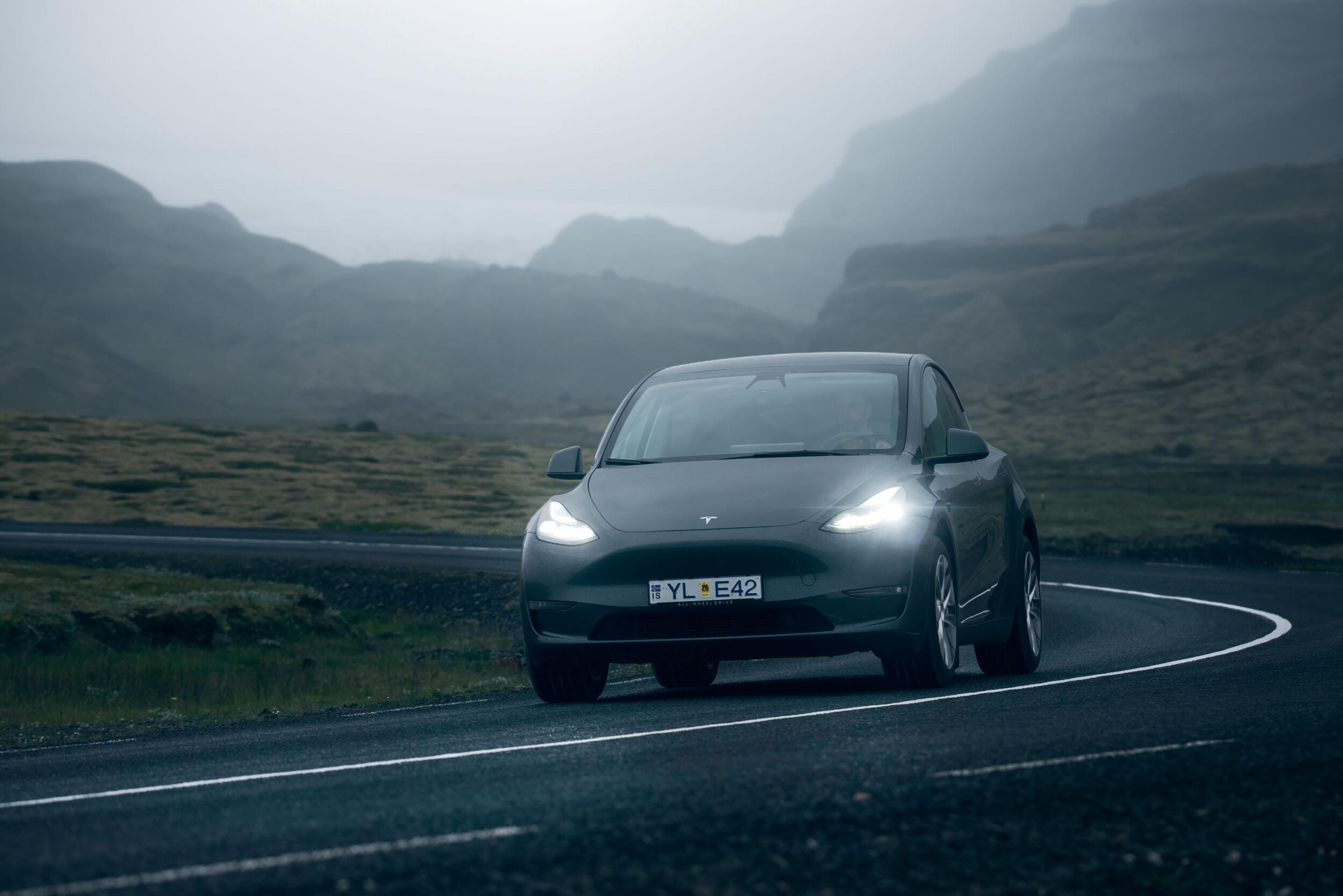  A sleek black rental car navigates a curving road with mist-covered mountains in Iceland, showcasing scenic car rental routes.