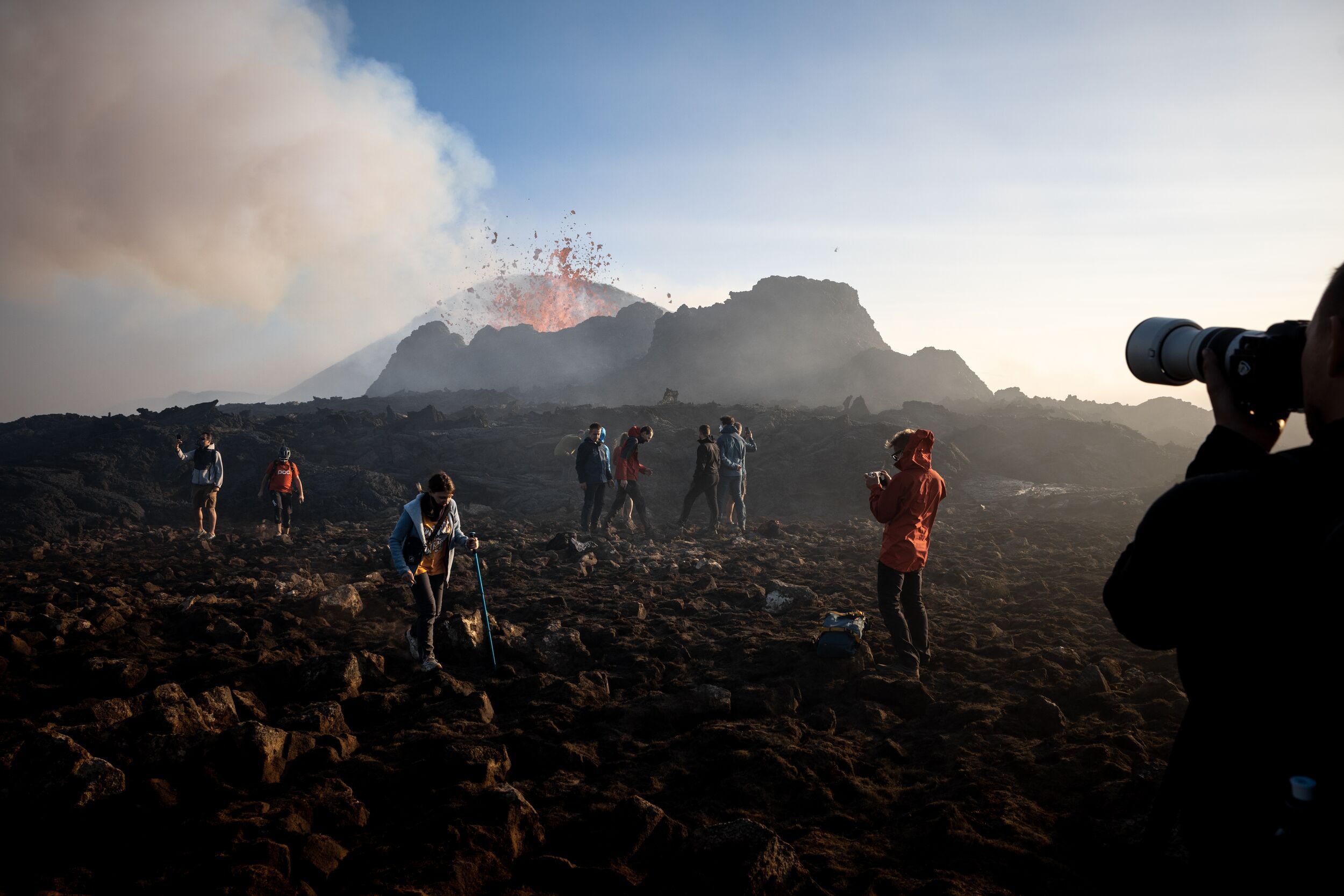Group of people photographing and observing a volcanic eruption on the Reykjanes Peninsula near Grindavik, with lava spewing from the crater and a rugged landscape in the foreground.