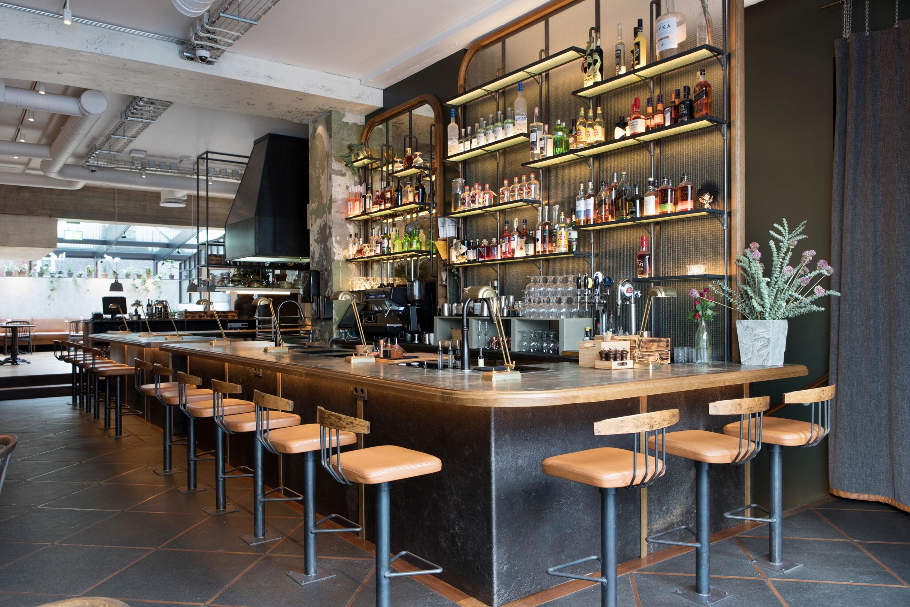 A modern, empty bar with a long counter, stools, and backlit shelves stocked with bottles.