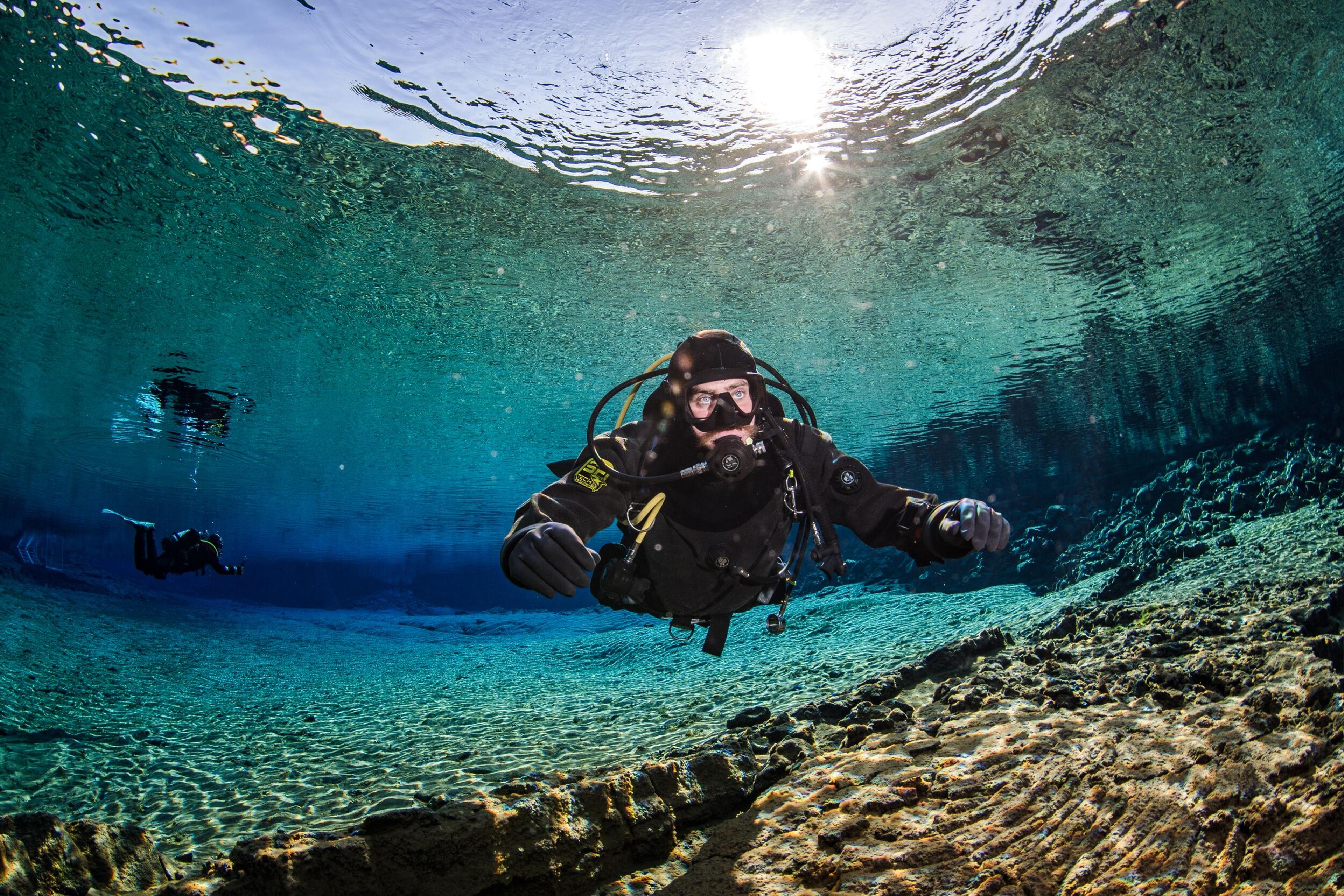 Scuba diver glides through crystal-clear water above rocky terrain, with sunlight reflecting on the water's surface.