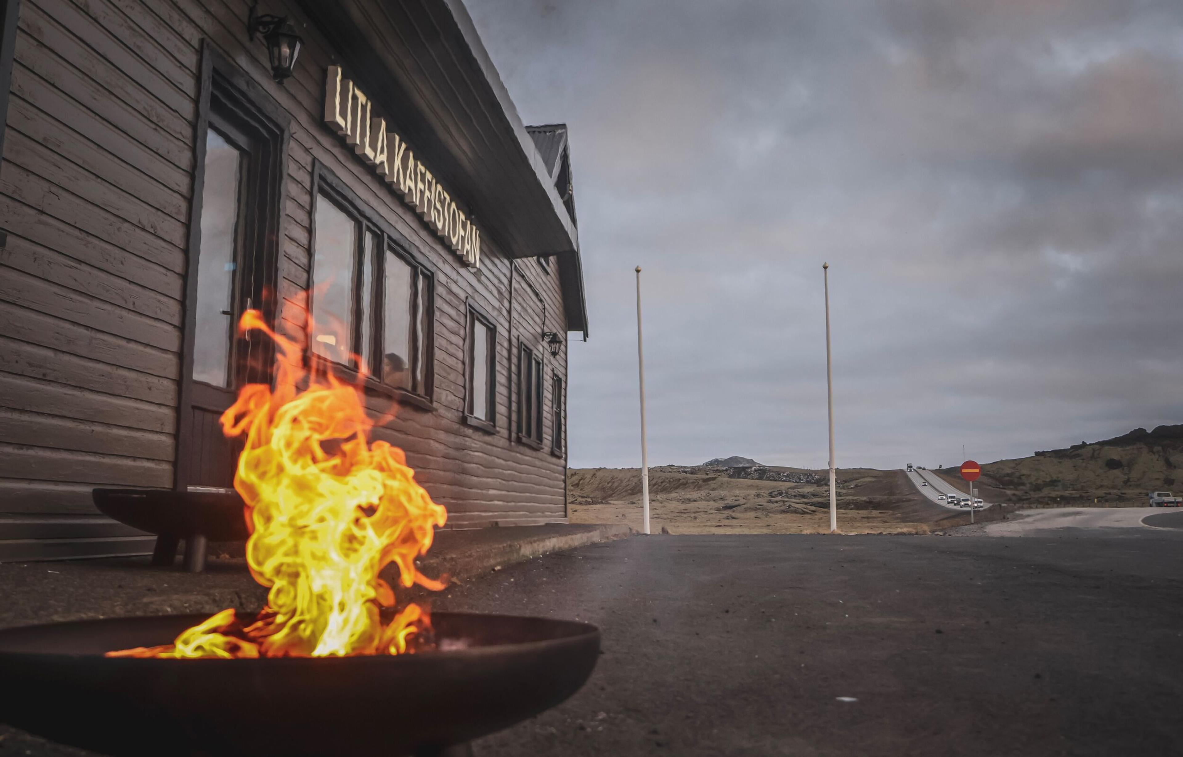 Bright fire in a pit in front of a wooden building named "Litla Kaffiskopan", with a desolate road and cloudy sky in the background.