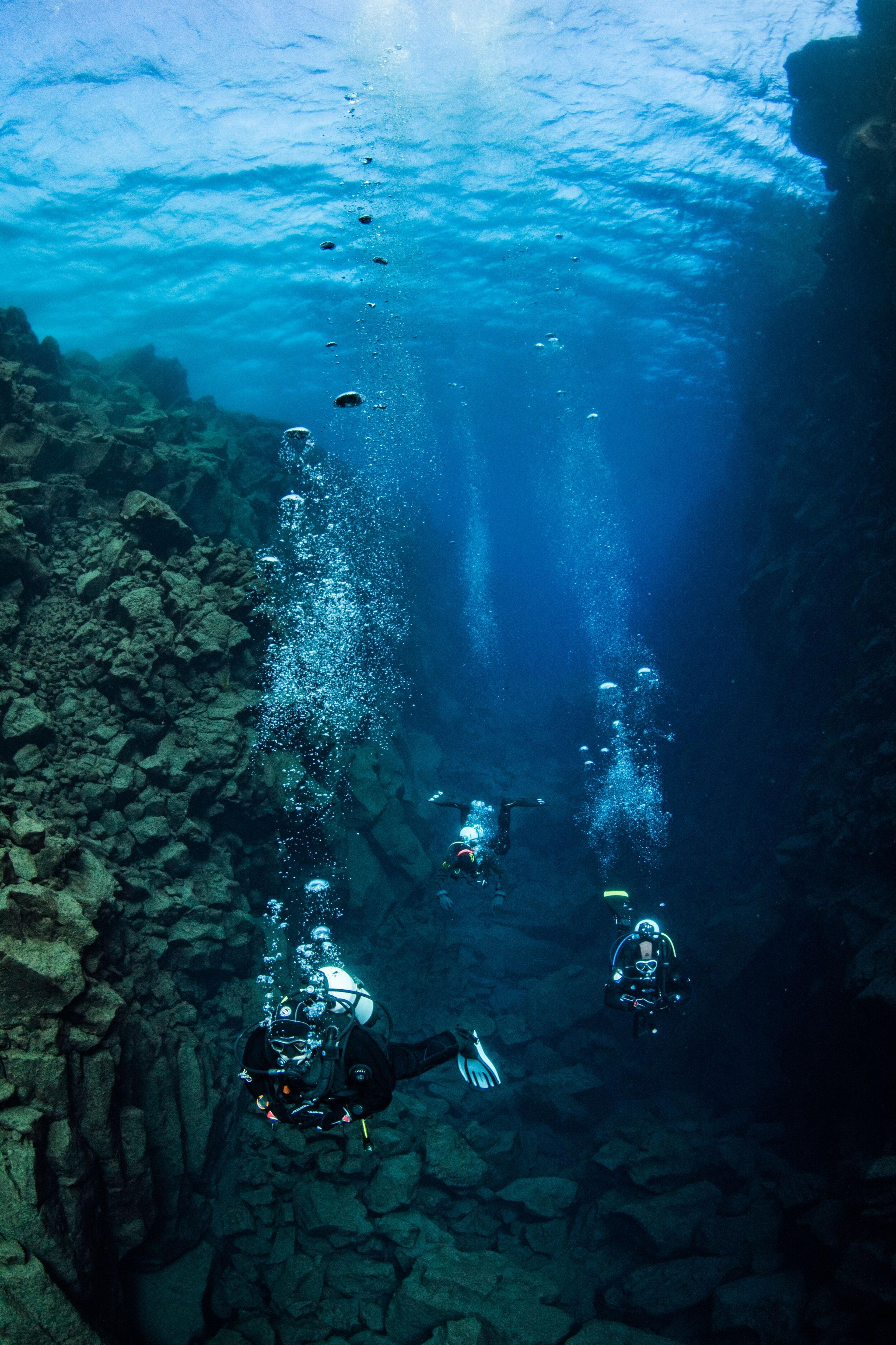 Three scuba divers in a deep, clear underwater fissure with rocky walls and rising bubbles.
