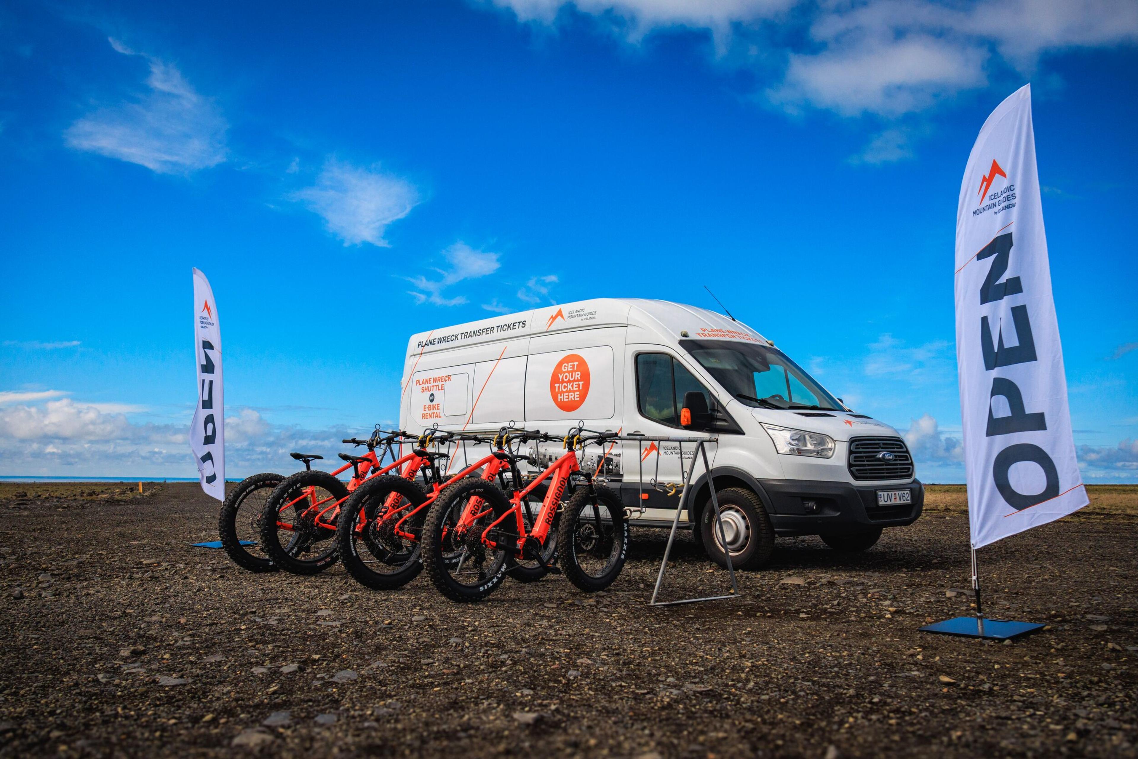 White van advertising "PLANE WRECK TRANSFER TICKETS" with several orange fat bikes in front, flanked by two "OPEN" flags, on a gravel ground under a bright blue sky.