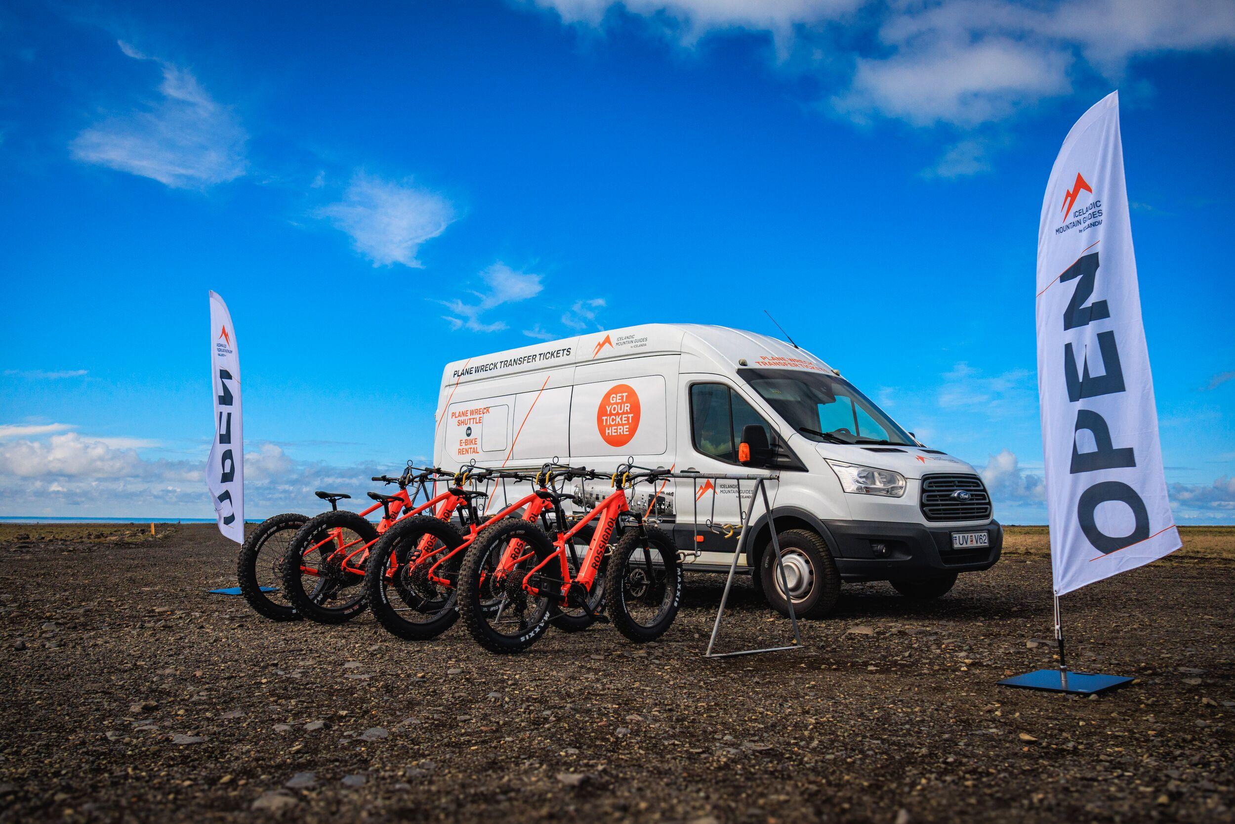 White van advertising "PLANE WRECK TRANSFER TICKETS" with several orange fat bikes in front, flanked by two "OPEN" flags, on a gravel ground under a bright blue sky.
