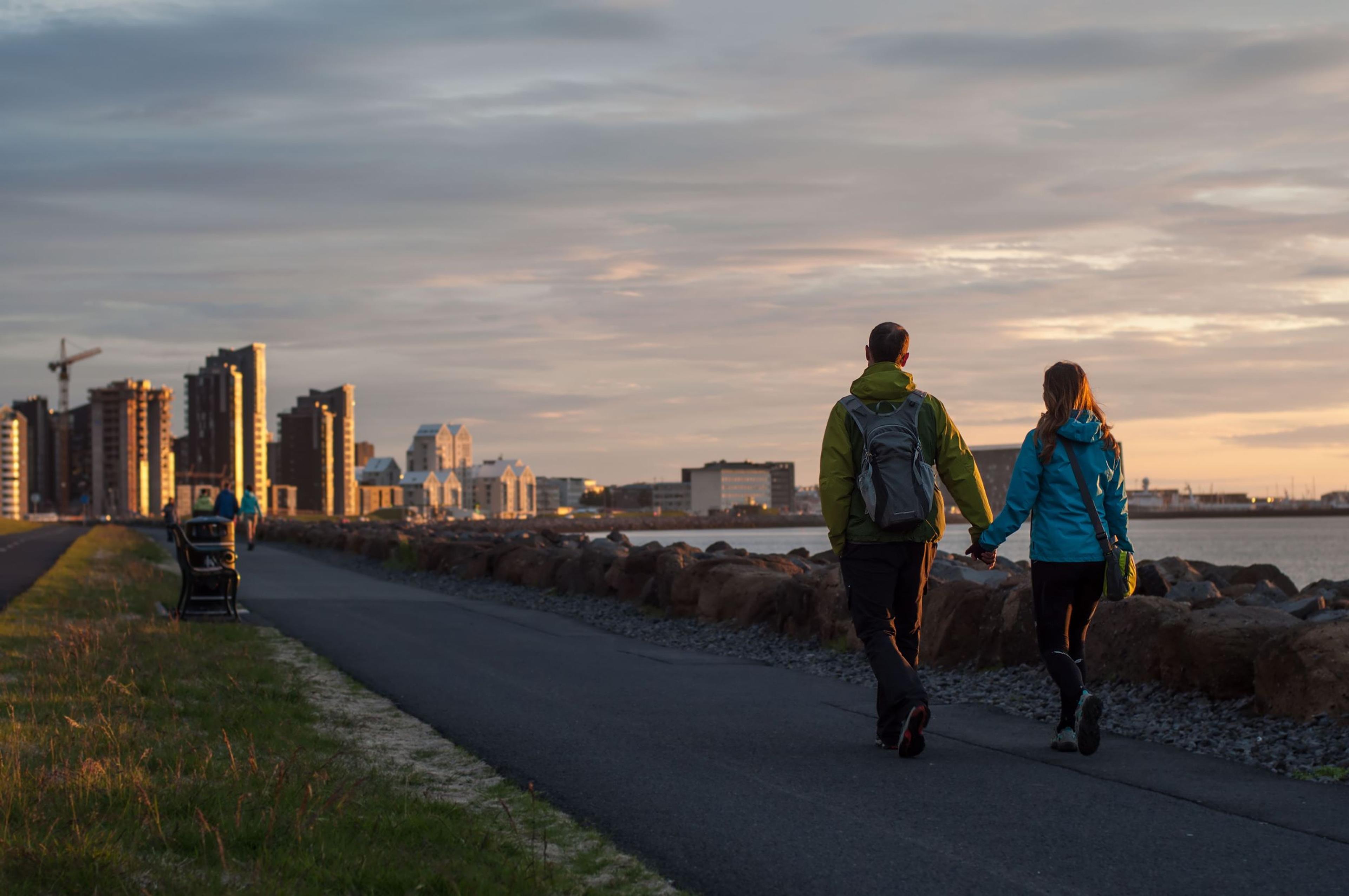A couple strolling along the coast of Reykjavík at sunset, with the city's tallest skyscrapers silhouetted in the background