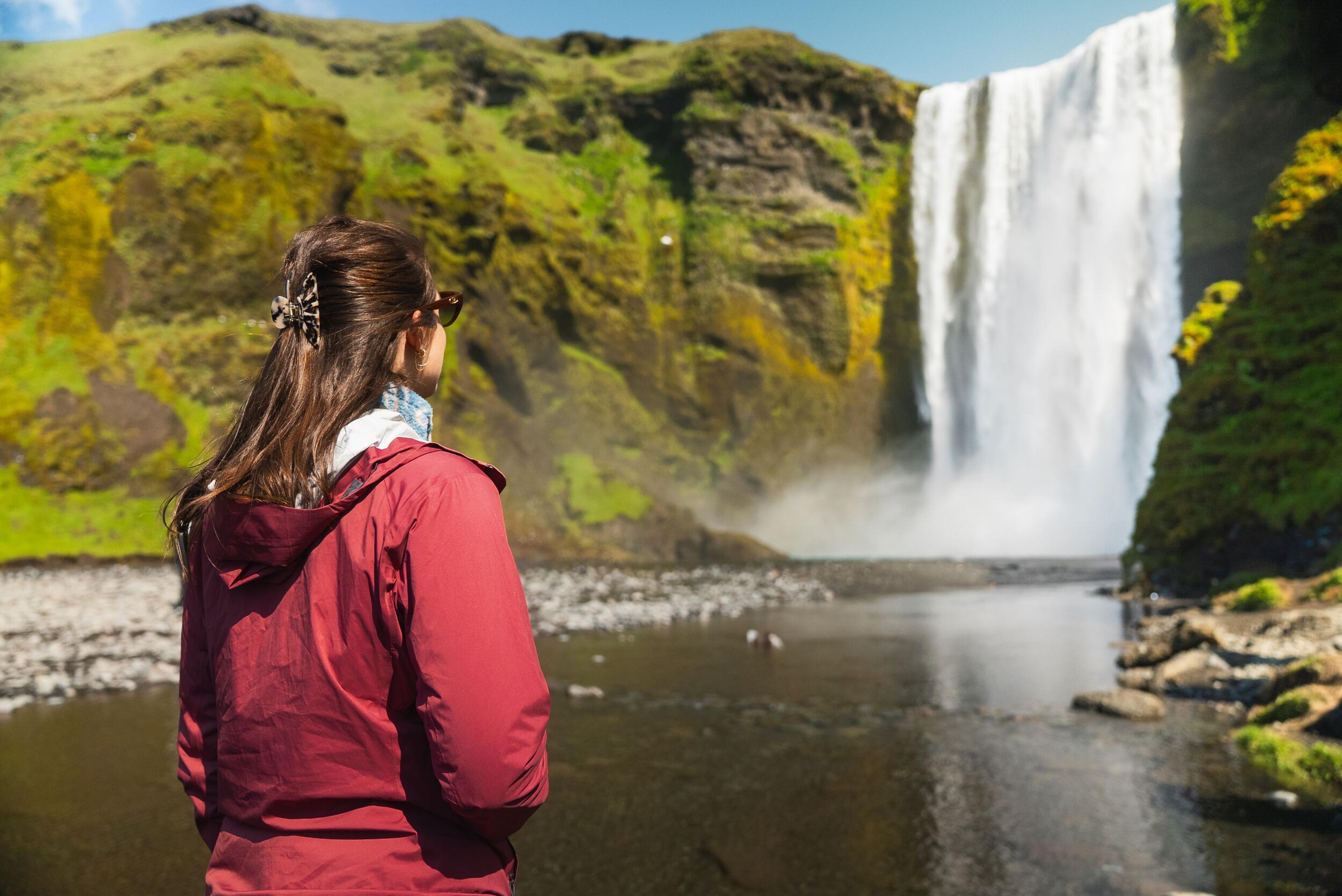 Person in a red jacket looking at a powerful waterfall surrounded by green, mossy cliffs.