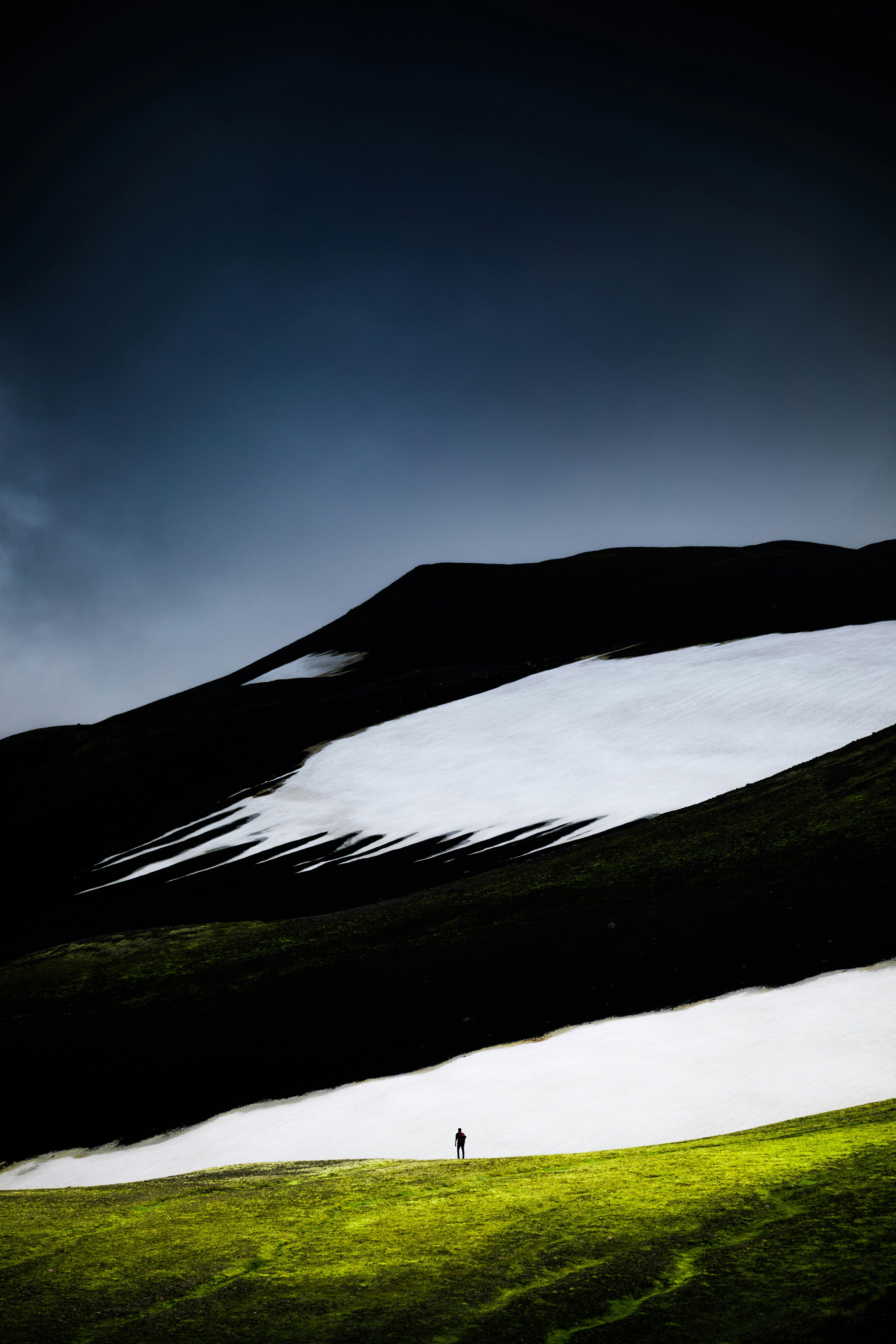 Lone hiker on a vibrant green field with contrasting dark hills and snow patches in the Icelandic Highlands at dusk.