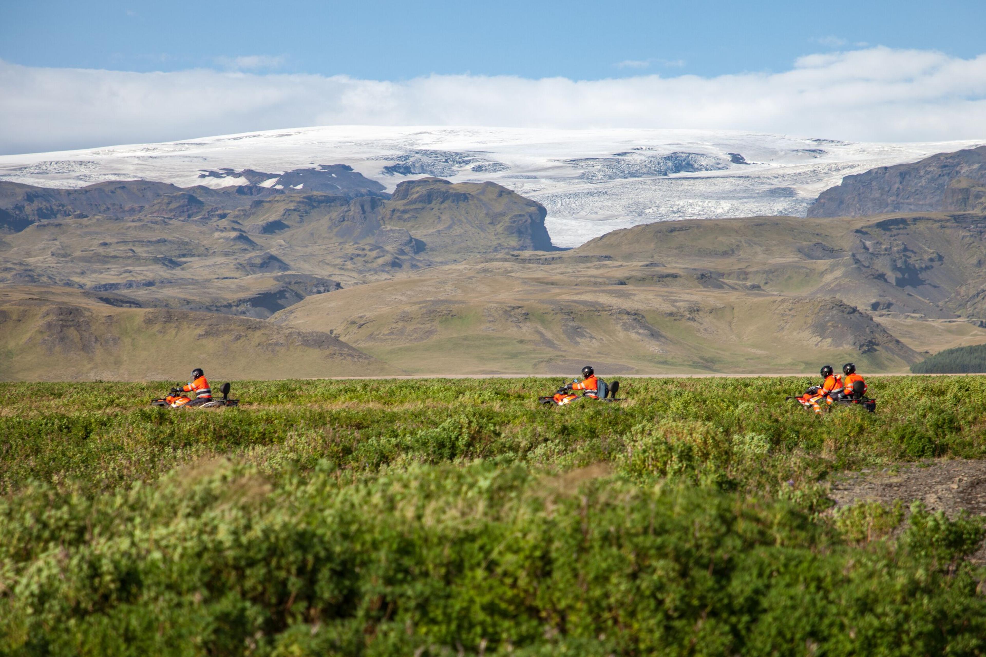 Three ATV riders in orange gear traverse a lush green valley, with towering mountains and a glacier-covered peak in the background, showcasing Iceland’s diverse landscapes.