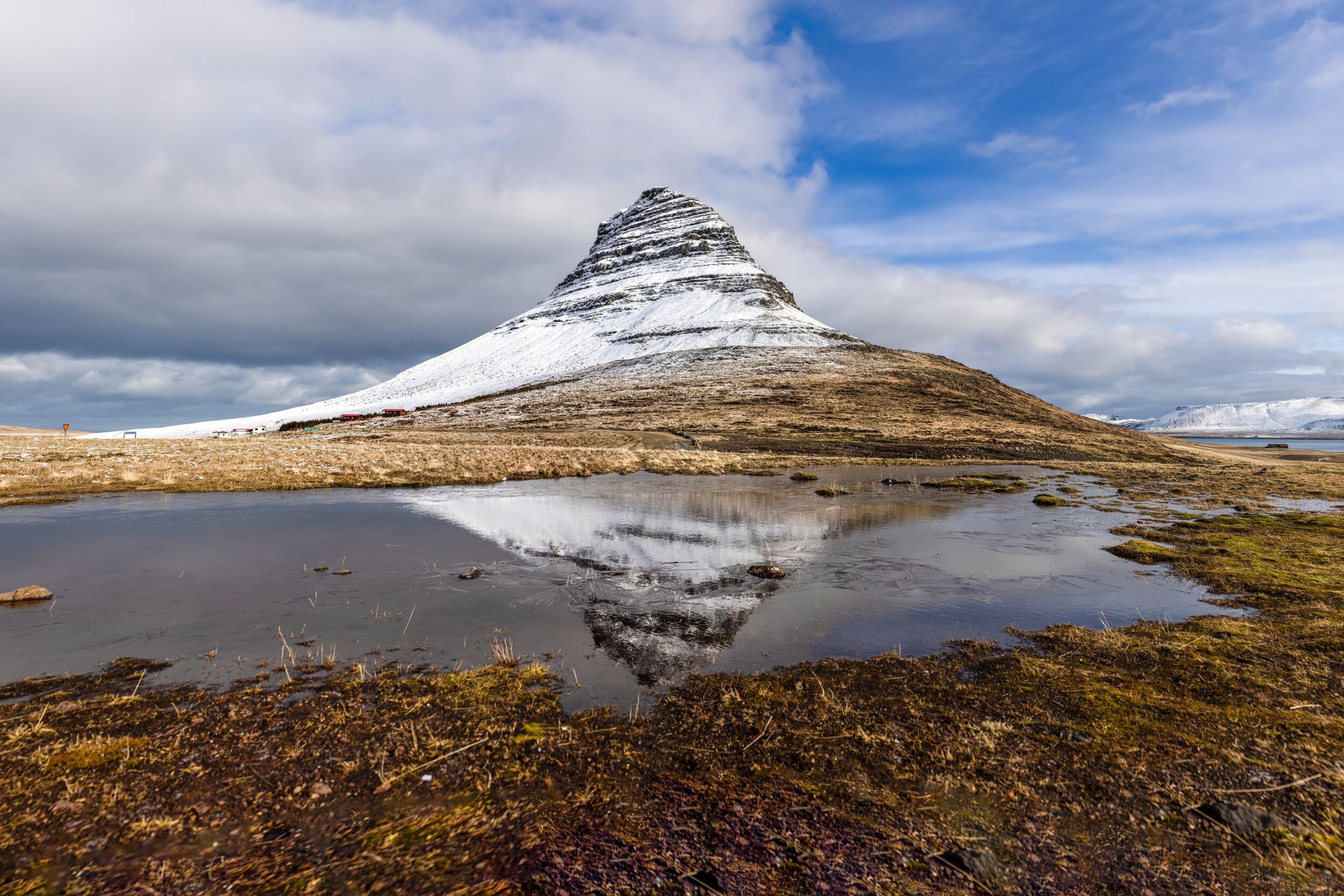 Snow-capped Kirkjufell mountain reflected in a still pond with grassy foreground under a partly cloudy sky.