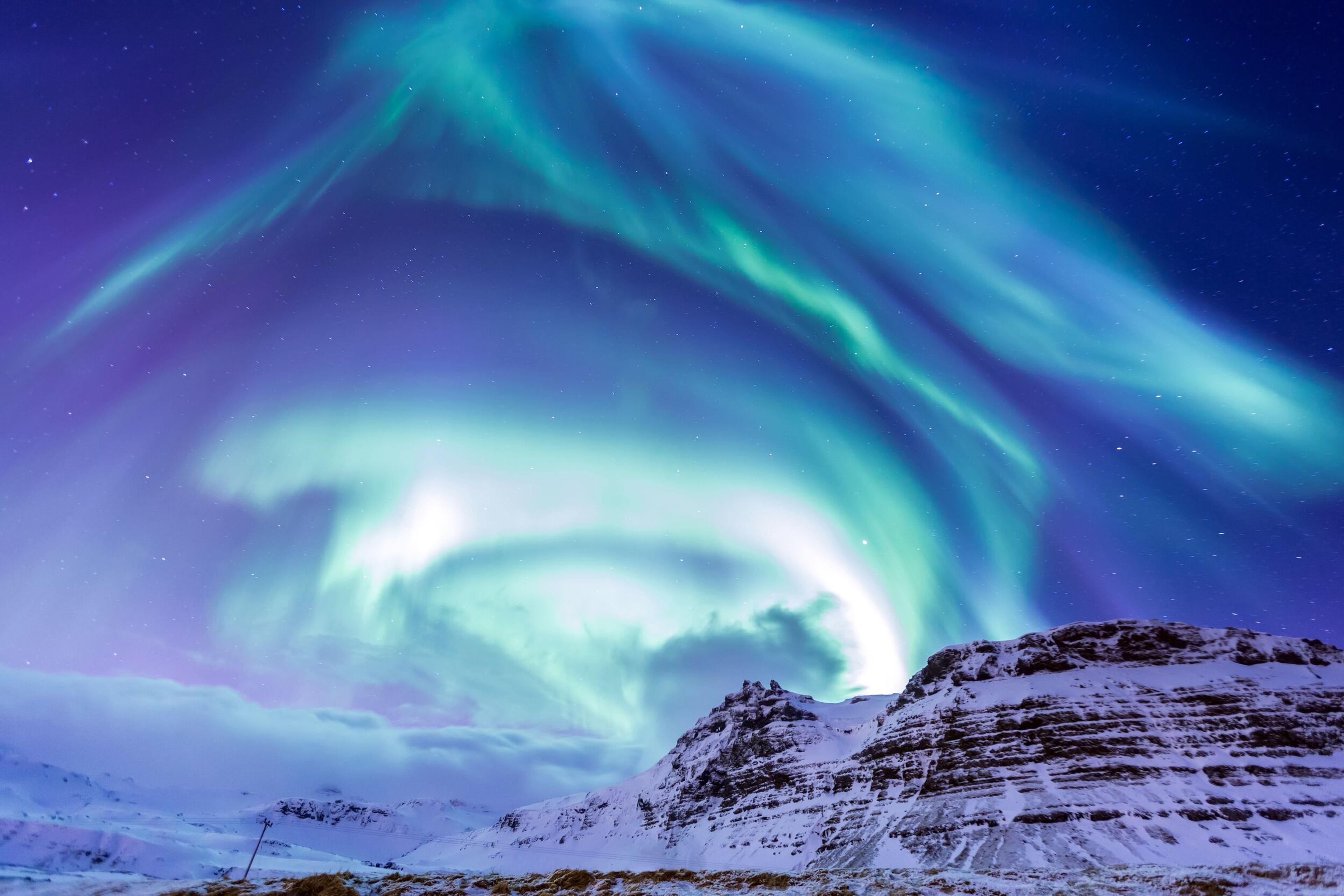 Vibrant green and purple aurora borealis swirl above snow-covered mountains.