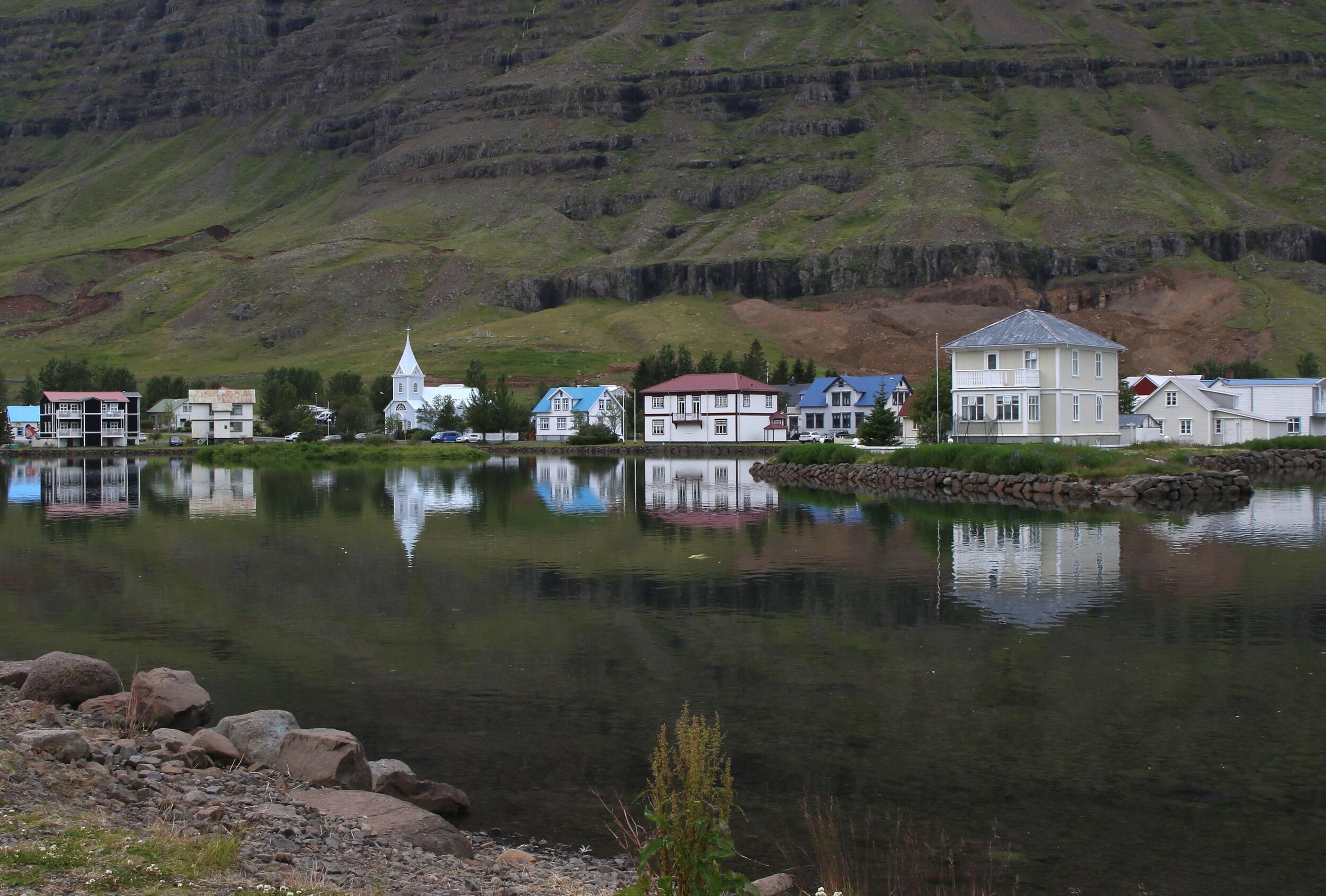 A small town with colorful houses and a white church reflected in calm water, set against a backdrop of green mountains.