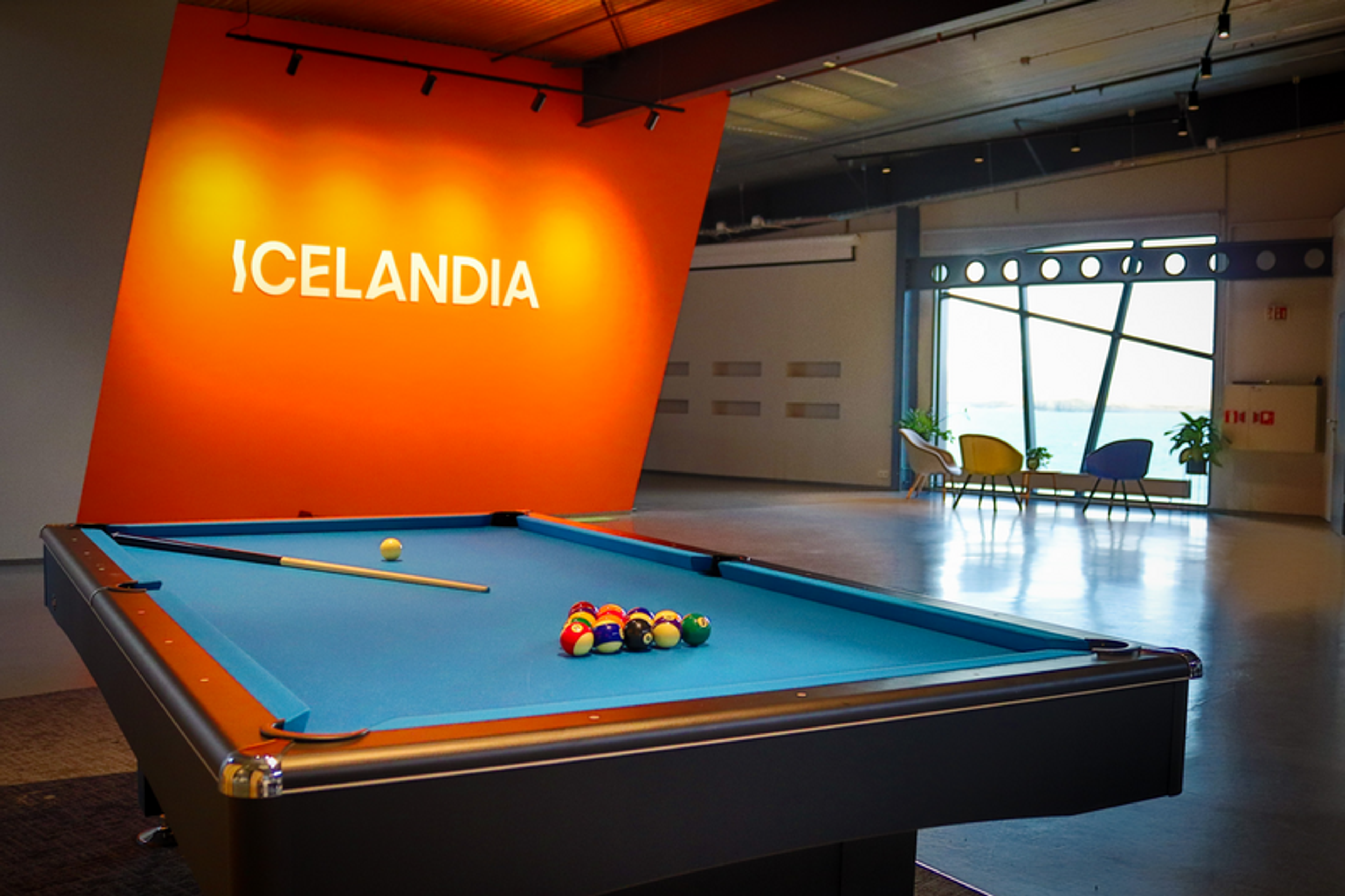  A recreational area with a pool table at the center, under overhead lighting. The wall is prominently branded with "ICELANDIA" in large, orange letters, giving the space a vibrant and energetic feel.