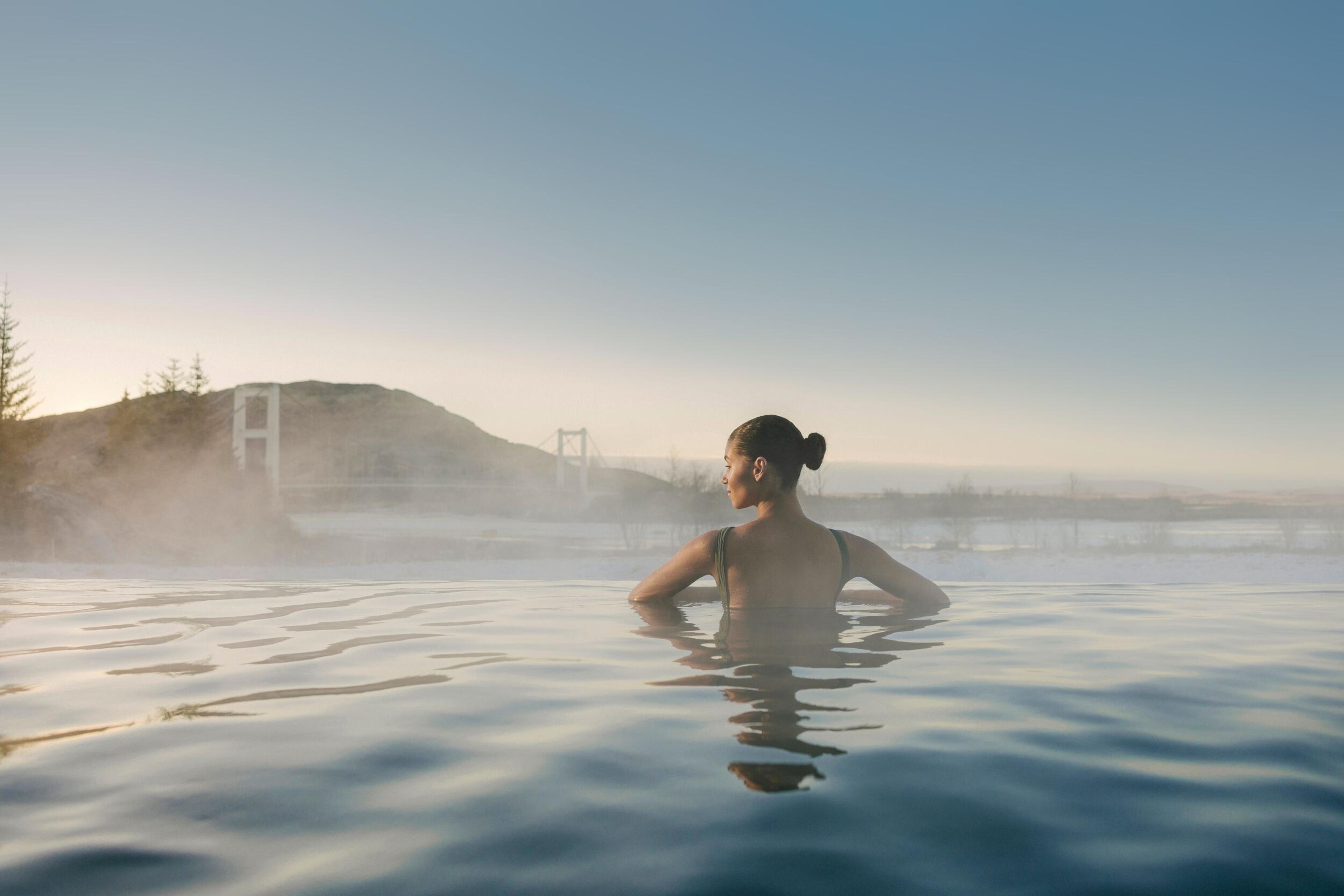 Woman in a steaming outdoor pool, looking towards a distant bridge and mountains.