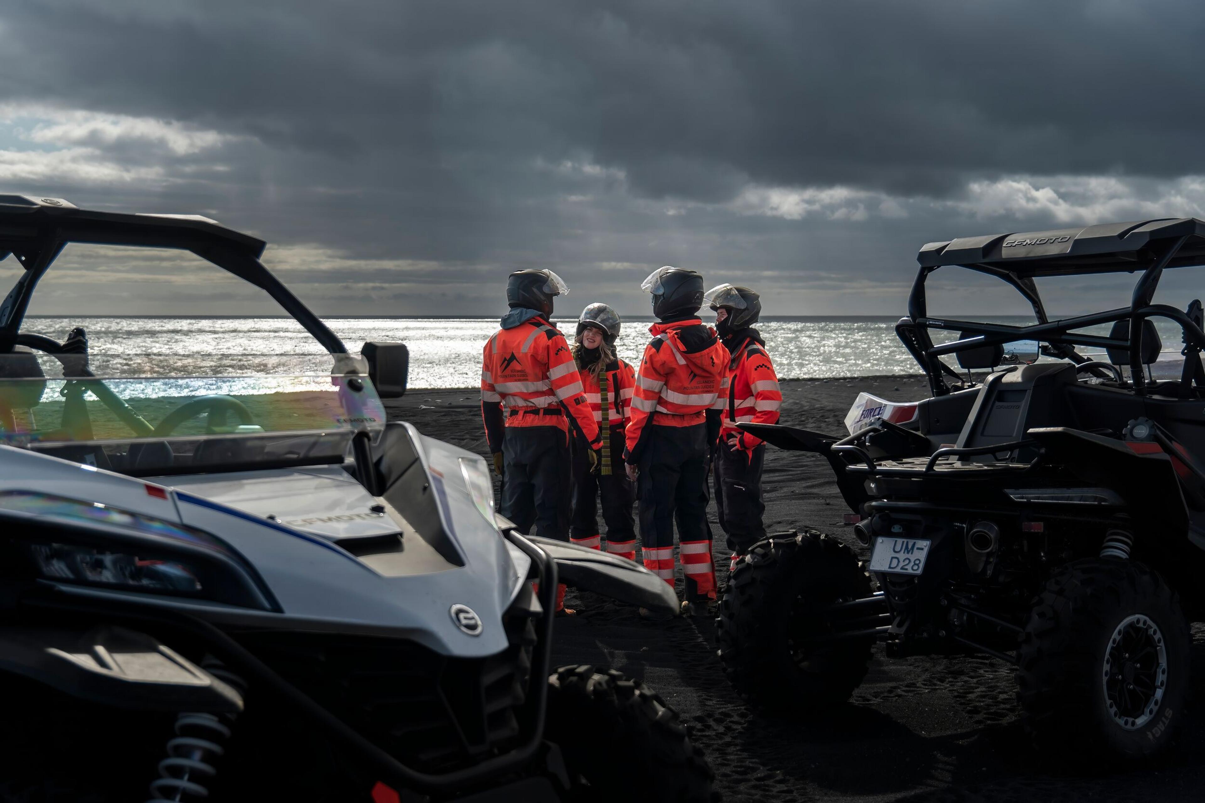 A group of ATV riders in high-visibility orange gear gathers near their parked off-road vehicles on a black sand beach, with the ocean and a dramatic sky in the background.