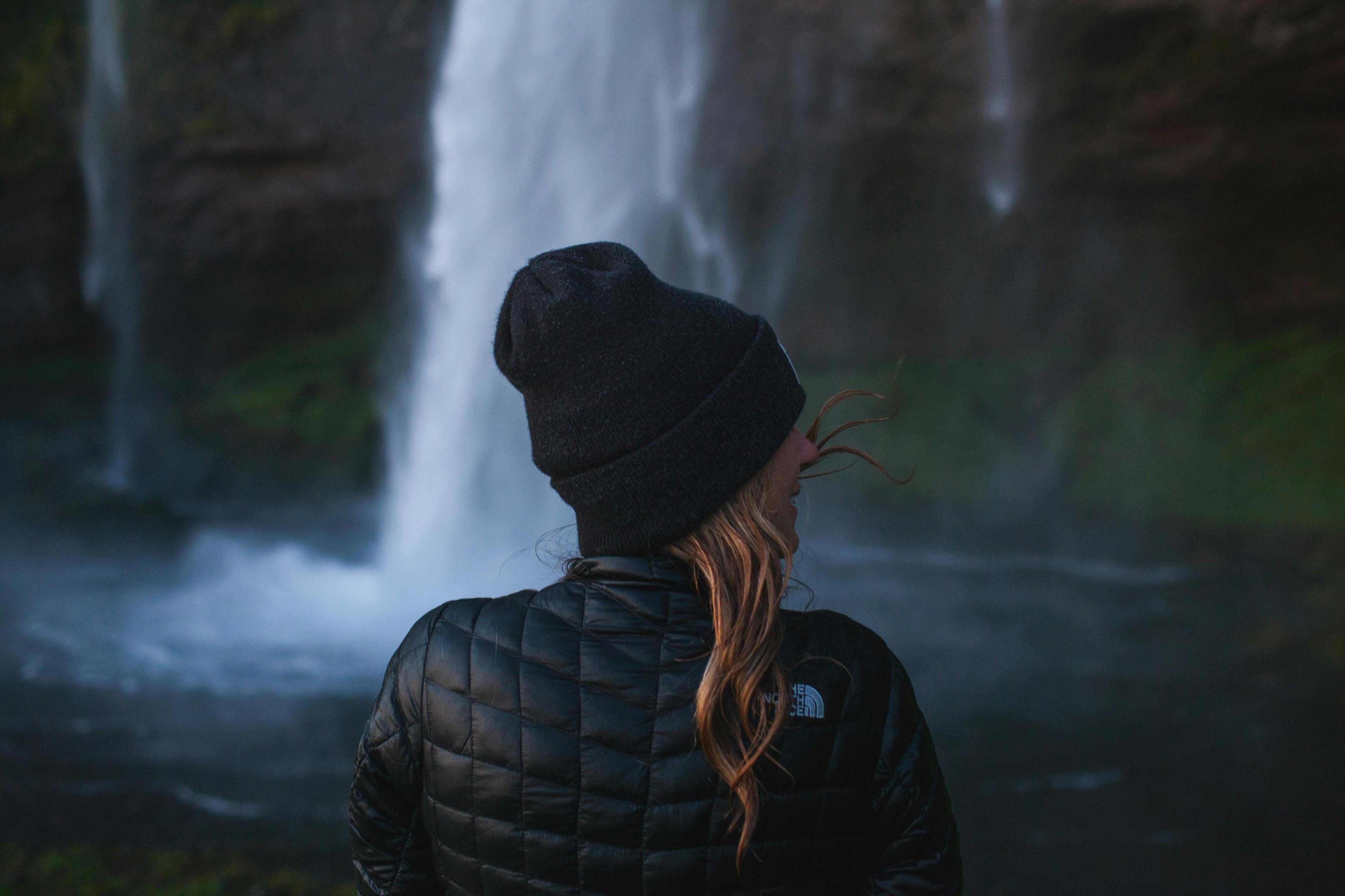 A woman in a black jacket and beanie gazes at the misty Seljalandsfoss waterfall cascading down in the background.