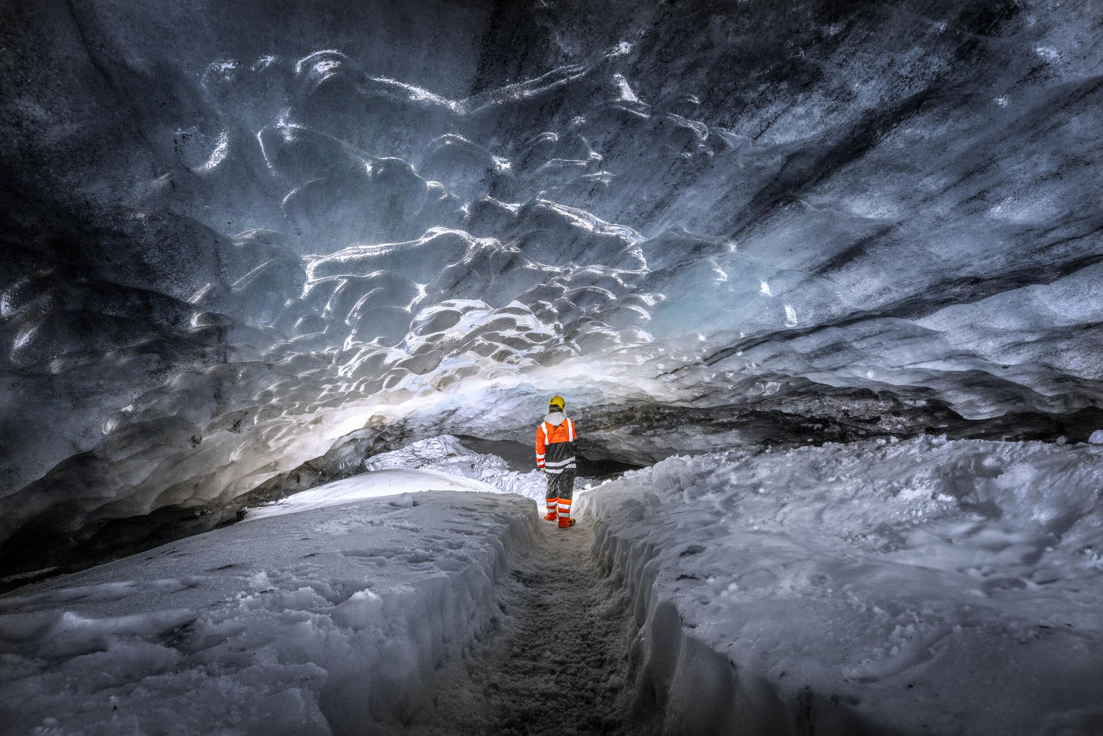 Explorer in orange and black suit pointing at the illuminated ice ceiling inside Askur ice cave, surrounded by snow.
