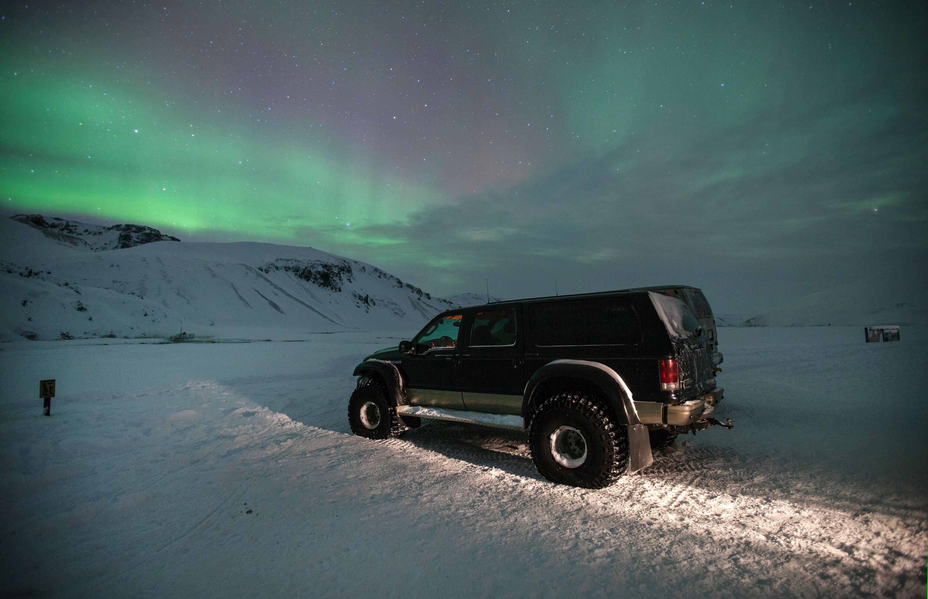 A large off-road vehicle is parked on a snowy landscape at night, with the green aurora borealis glowing in the sky above and snow-covered mountains in the background.