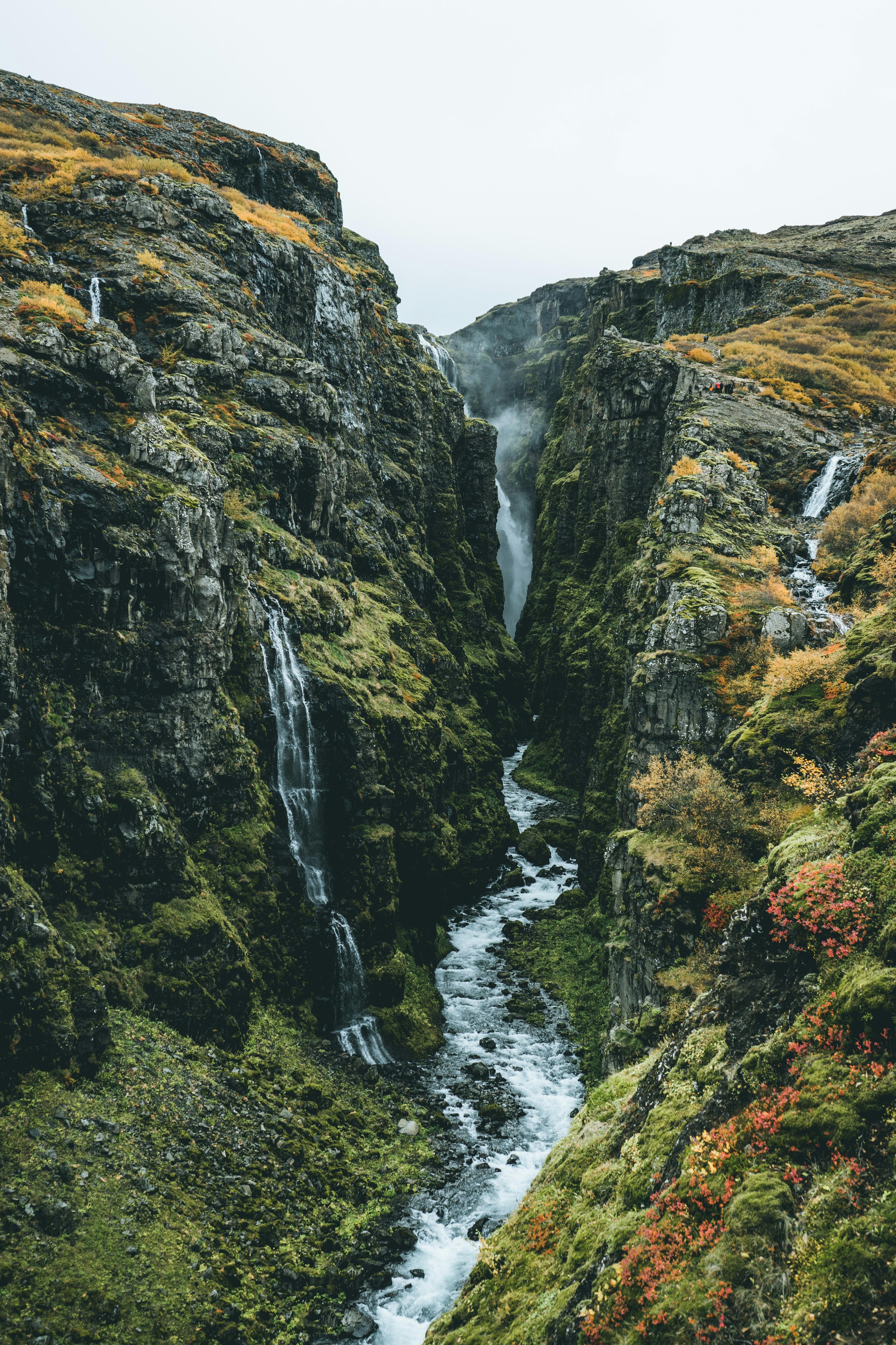 A narrow, verdant canyon dotted with small waterfalls cascading down its rocky cliffs, with a river flowing through the gorge surrounded by autumn-colored foliage.