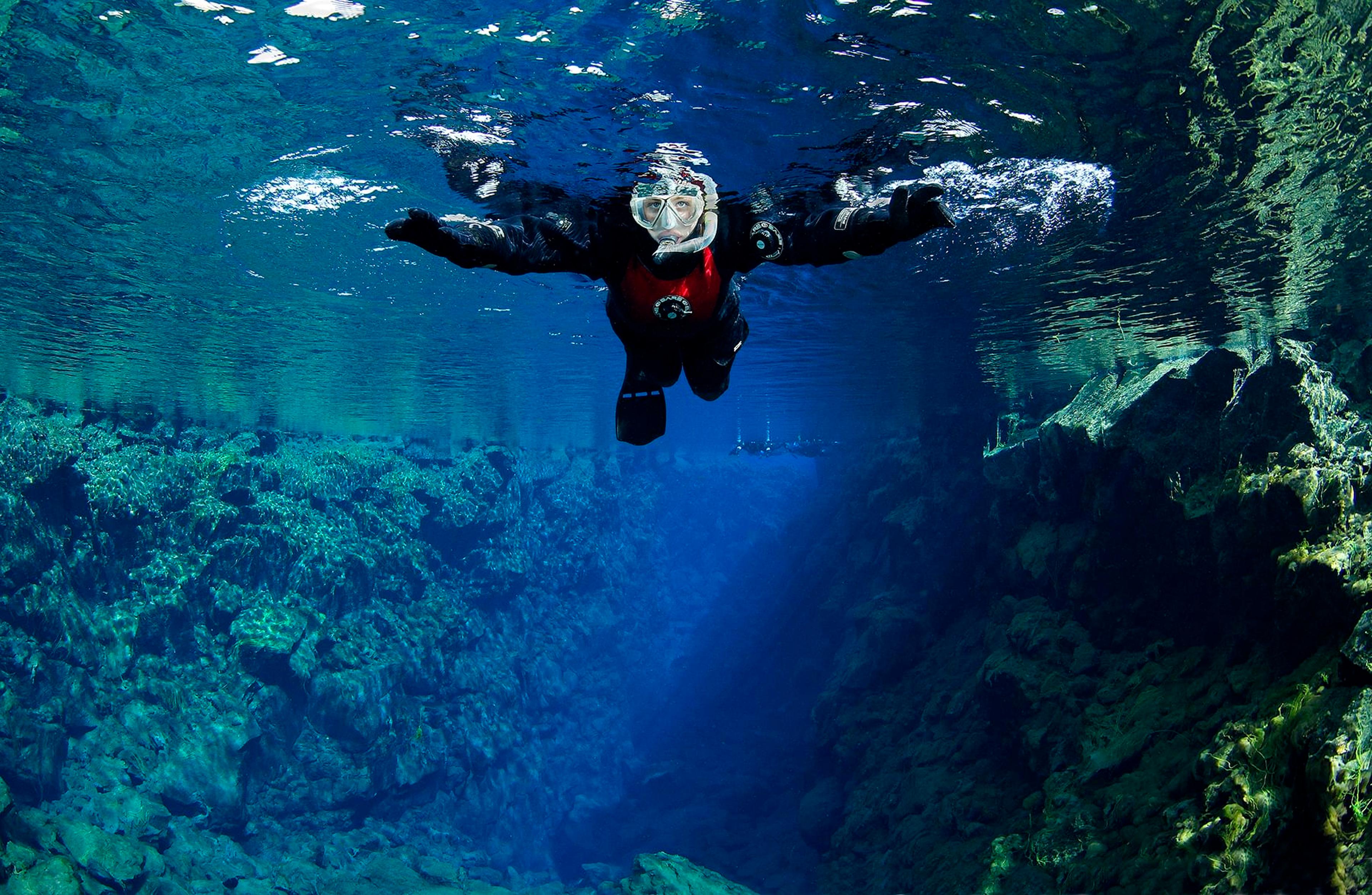 A person in a dry suit snorkeling in a clear blue underwater canyon.