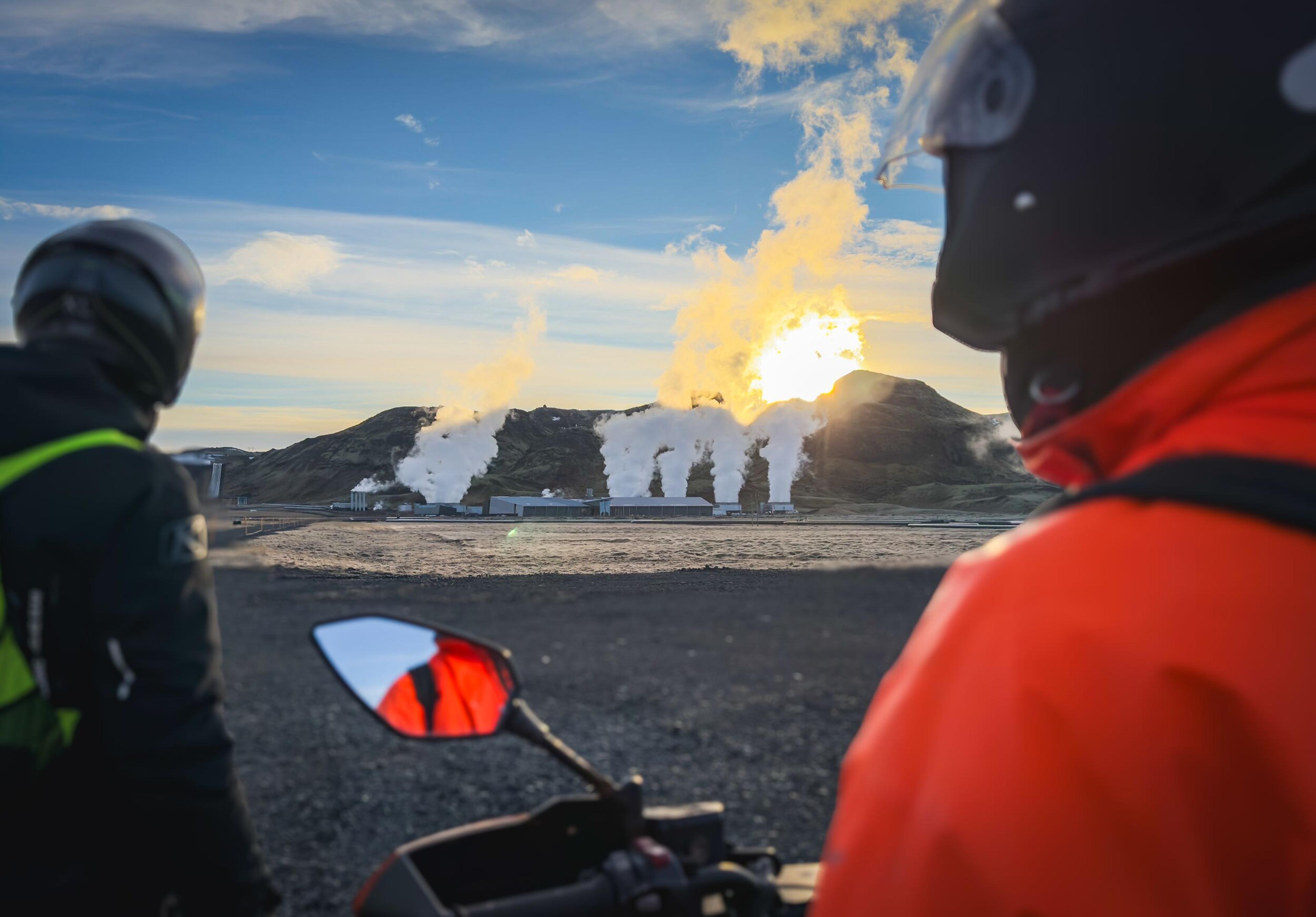 Two motorcyclists view a geothermal power plant with steam rising against a bright sun over hills.