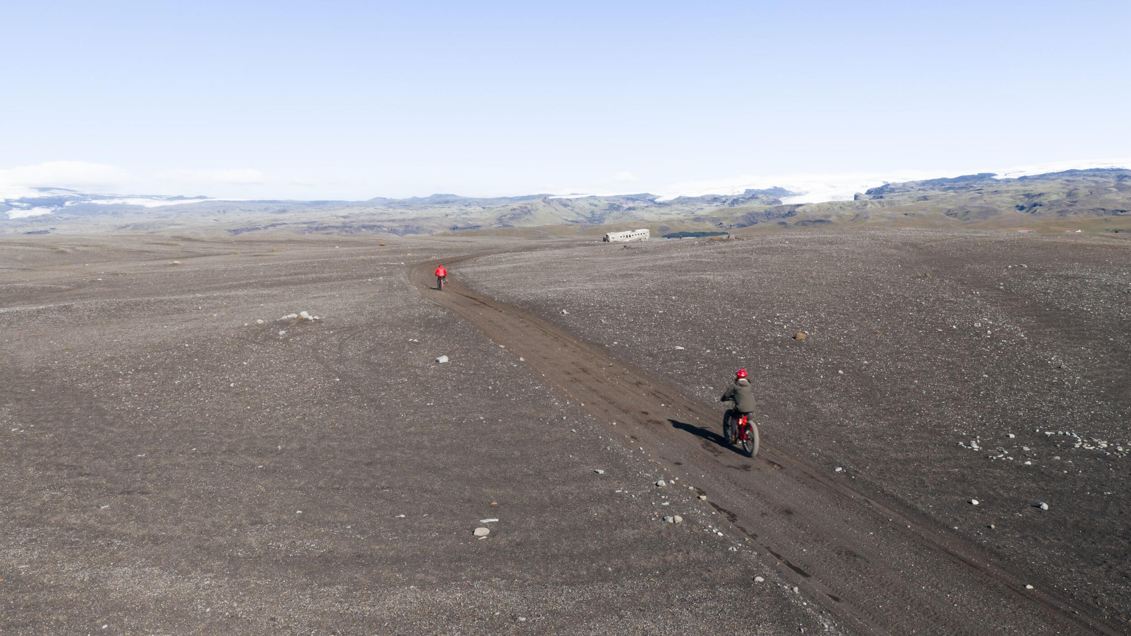 Two cyclists ride on a dirt road through a vast, barren, dark landscape under a clear sky.
