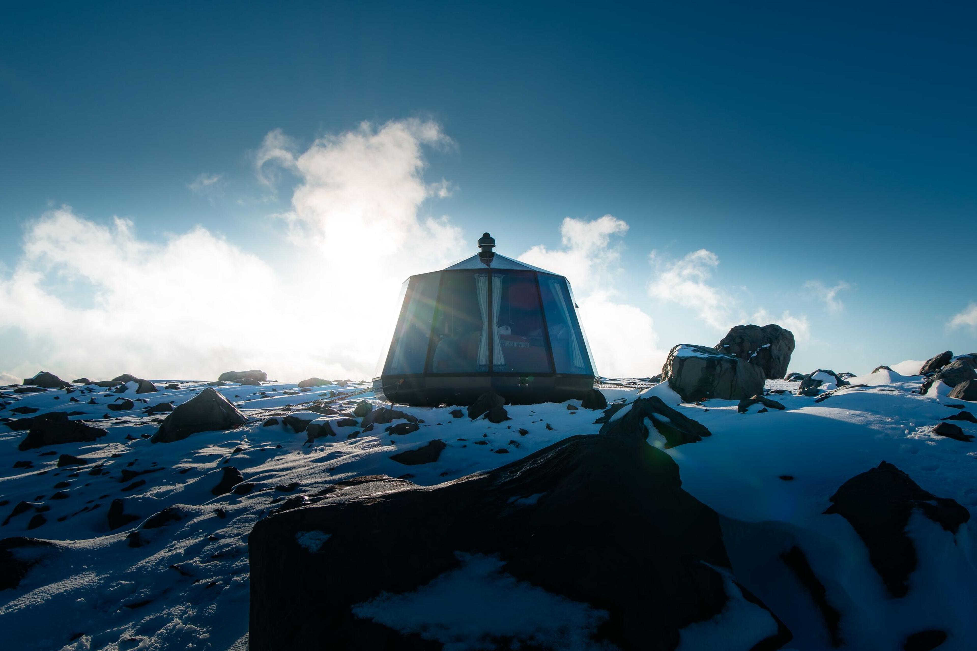 A futuristic glass-domed shelter on a snowy, rocky mountaintop under a bright sky.
