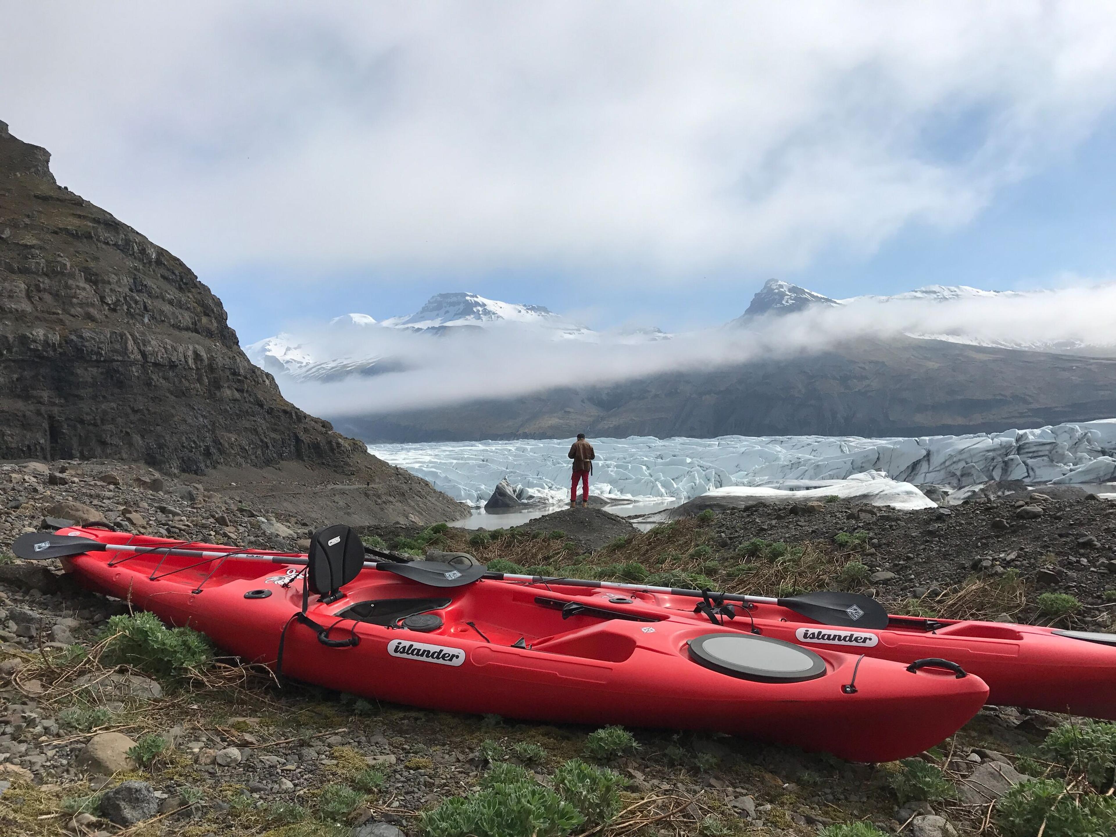 Two red kayaks rest on a rocky shore with a person standing in the background, overlooking a glacier and mountains, perfect for kayaking in Iceland.