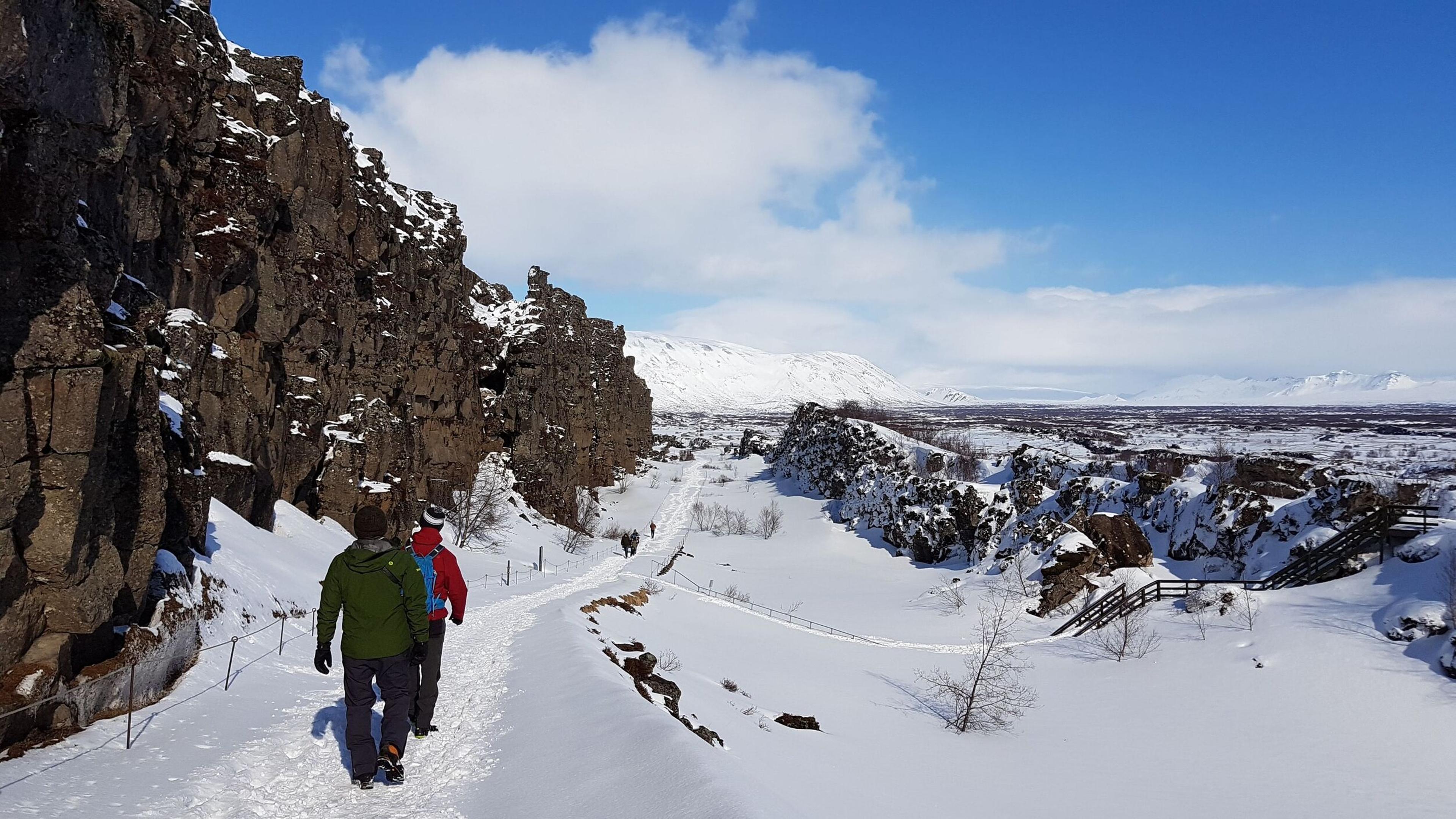 Two people walk on a snowy path between tall rock formations and a snow-covered valley with distant mountains under a bright blue sky.