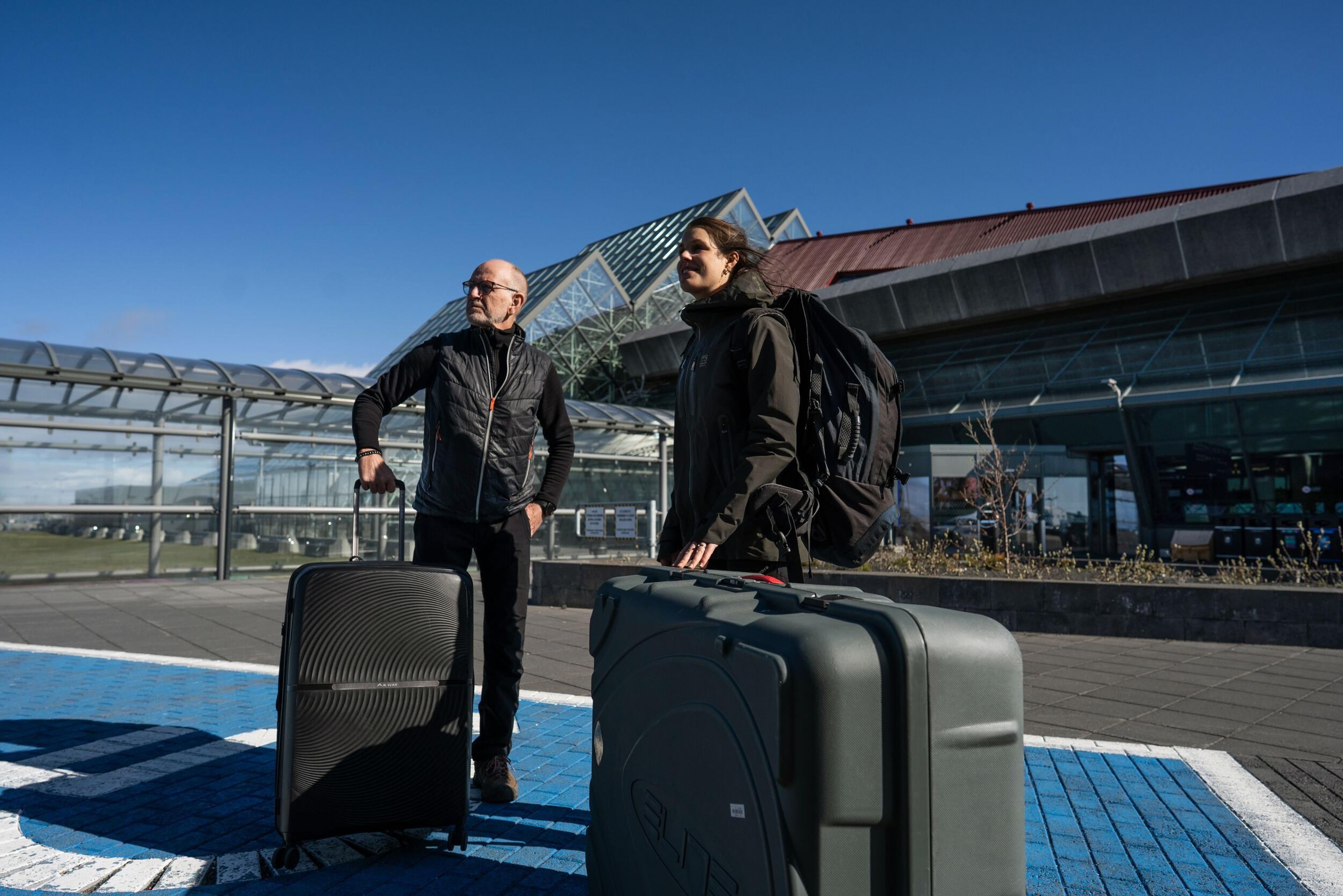 Two travelers with luggage, including a large hard case, stand outside an airport.