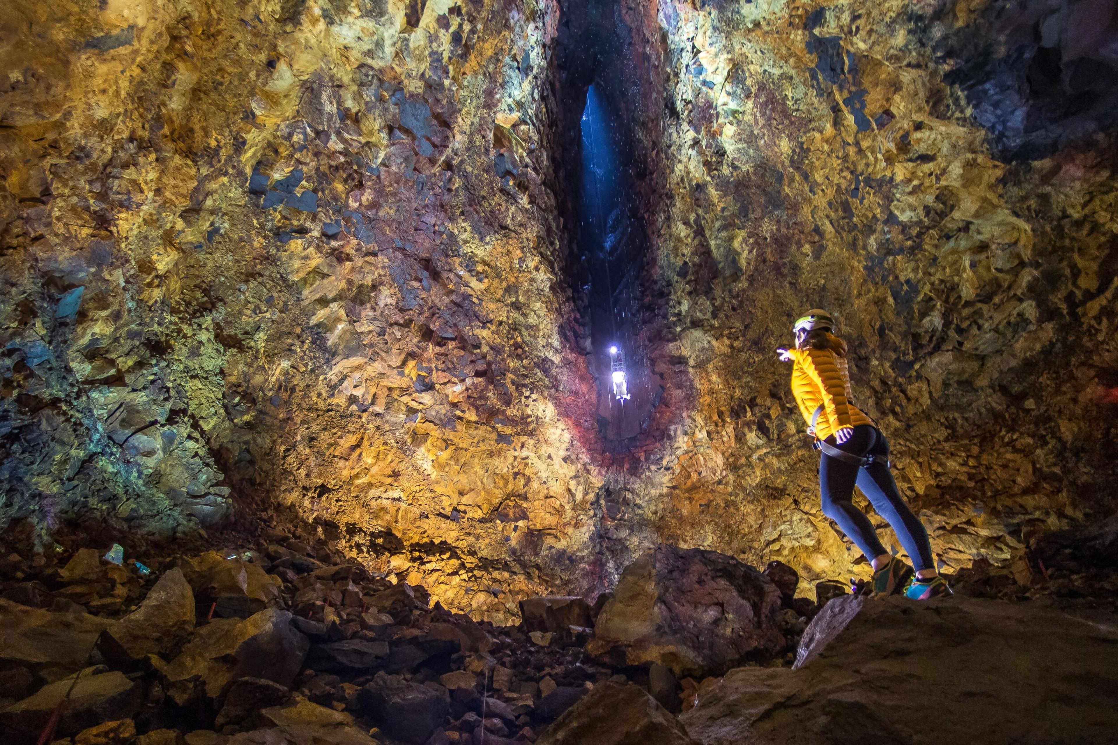 An adventurer in a yellow jacket points up a deep, colorful volcanic shaft where another person rappels.