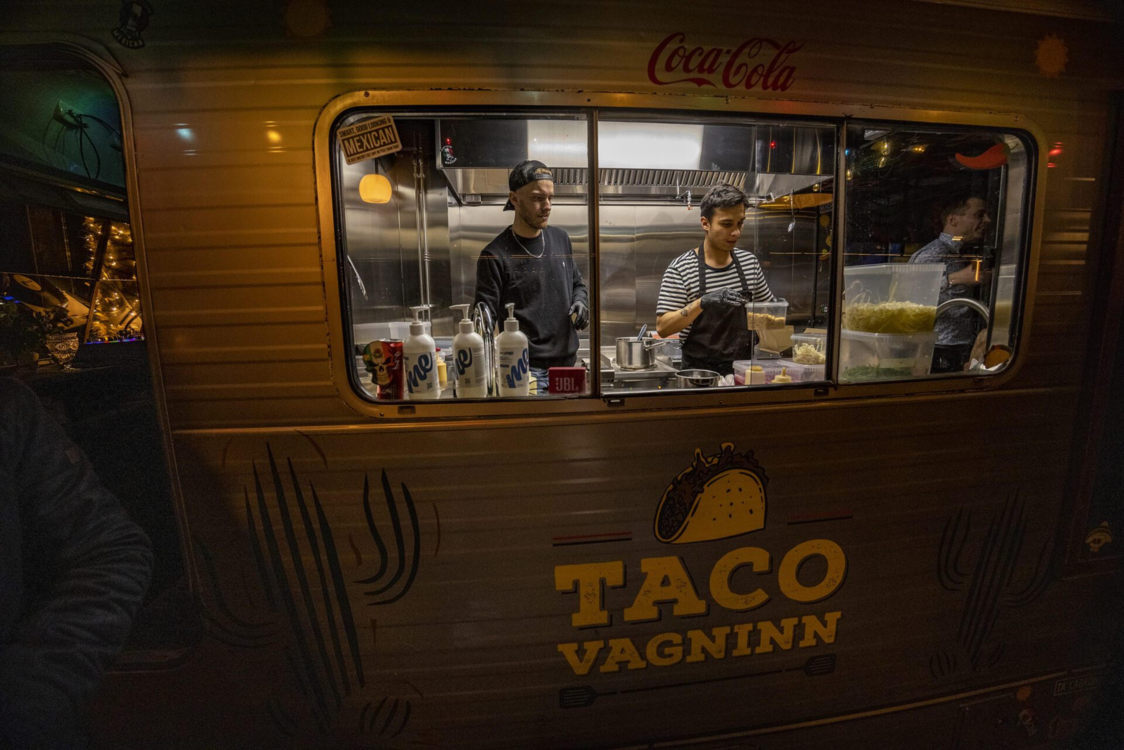 Chefs preparing food inside the Taco Vagninn food truck at The Greenhouse Food Hall in Hveragerdi, seen through the window of the truck with warm lighting.