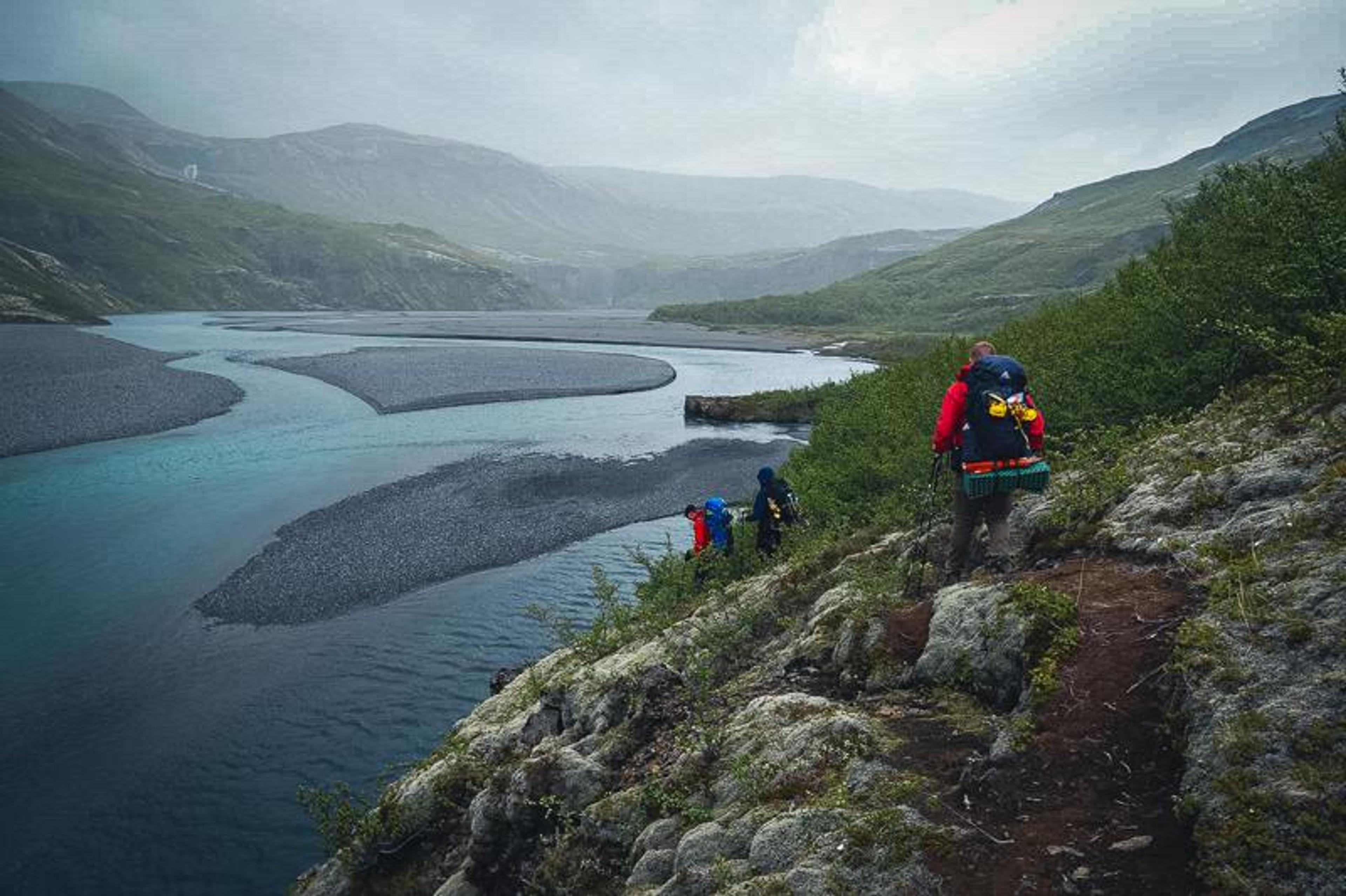 Three hikers on a rocky trail beside a wide river with gravel bars, surrounded by mountains under a cloudy sky.