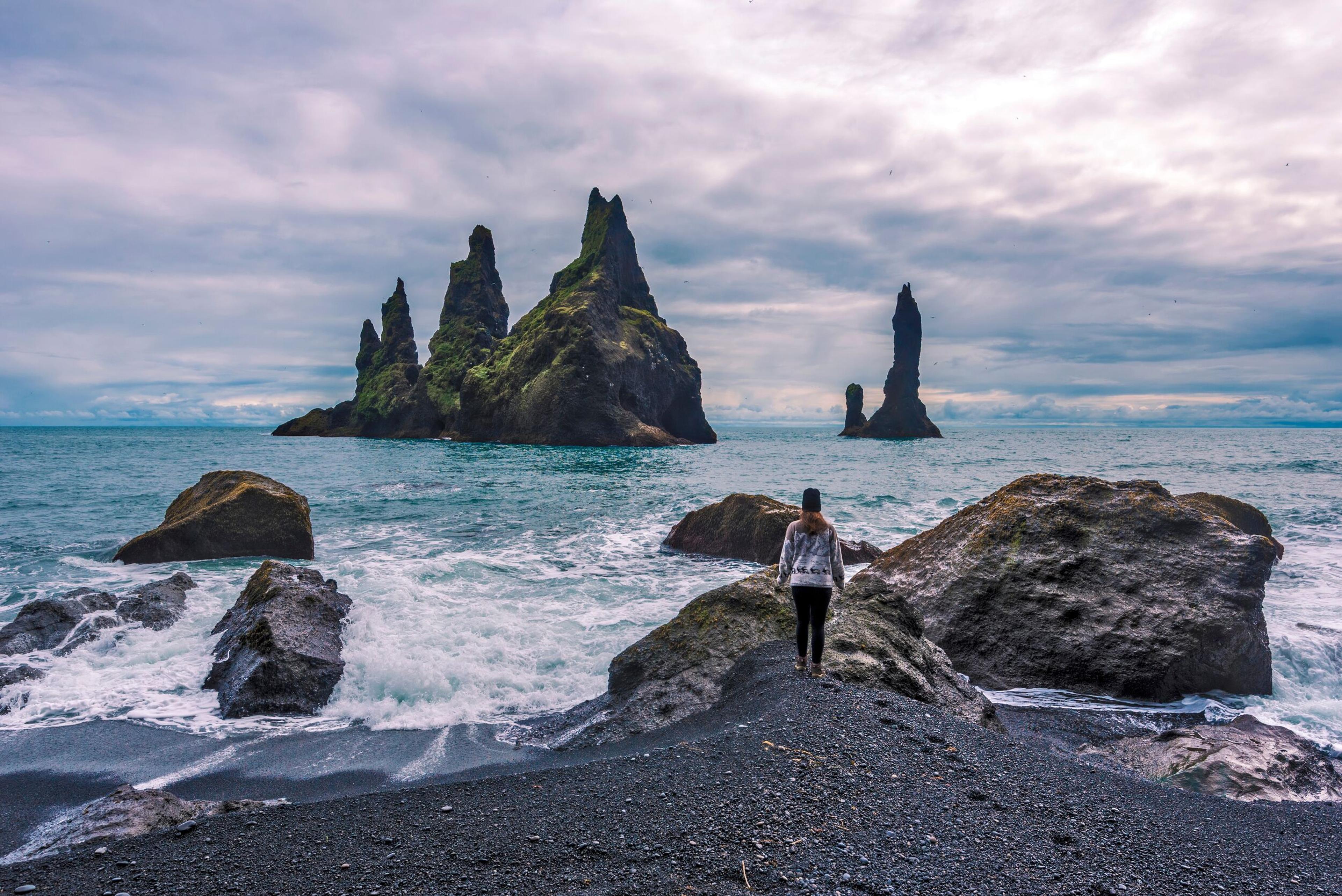 A person stands on a black sand beach looking at towering sea stacks in the ocean under a cloudy sky.
