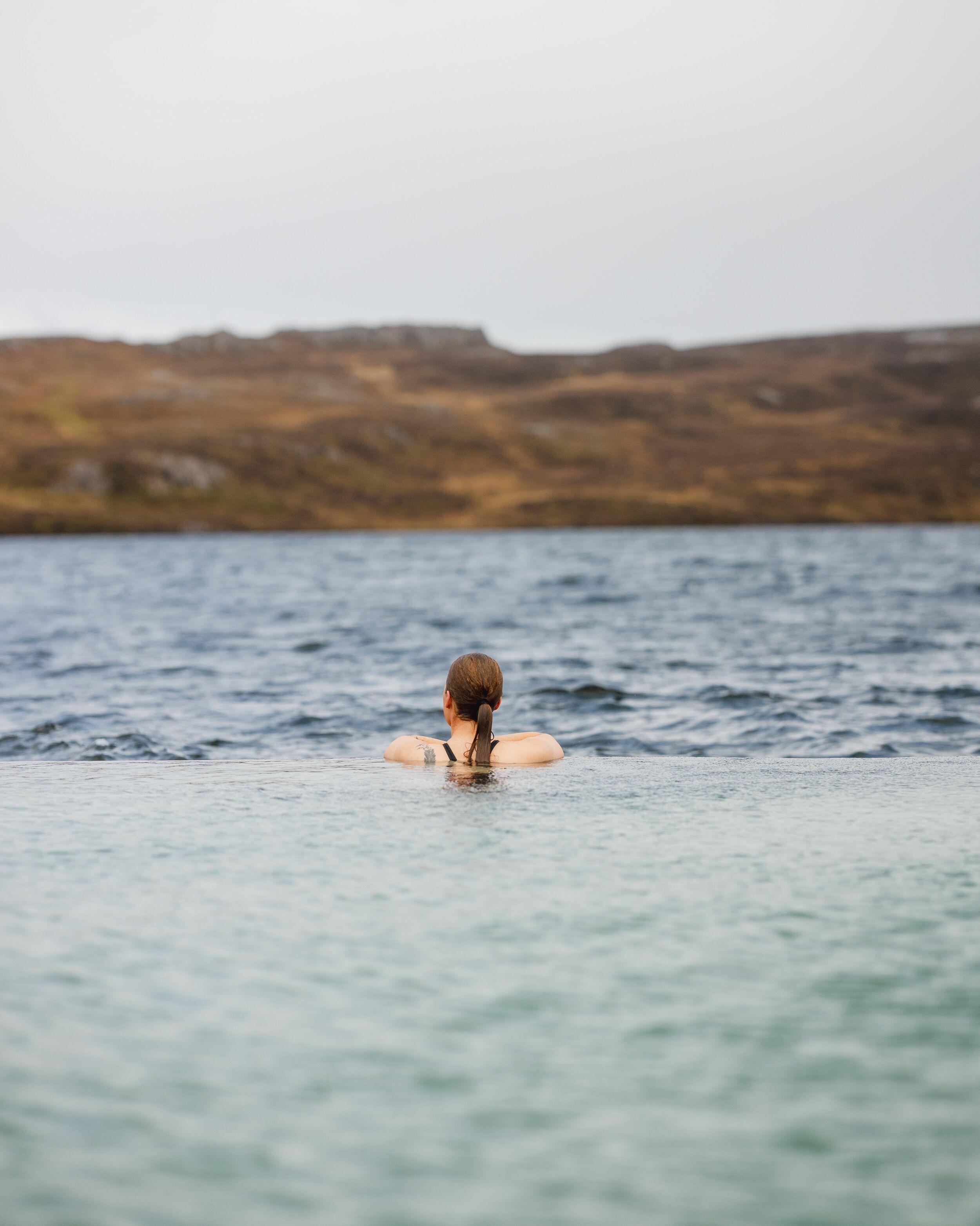 A person in an infinity pool looking out over a lake and distant brown hills under a grey sky.